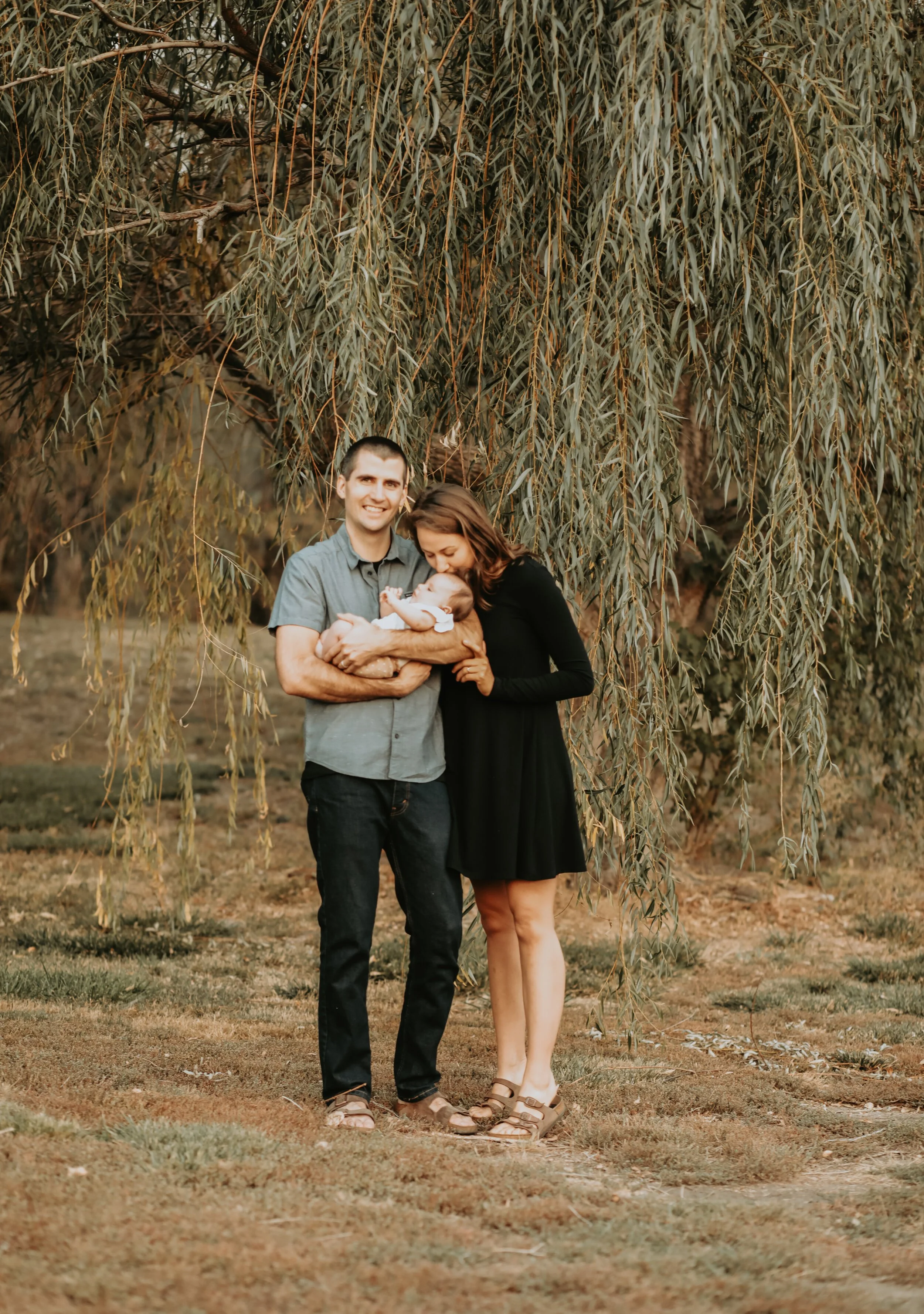 A family of three standing outdoors under a large hanging tree, with a man holding a baby and a woman kissing the baby's head.