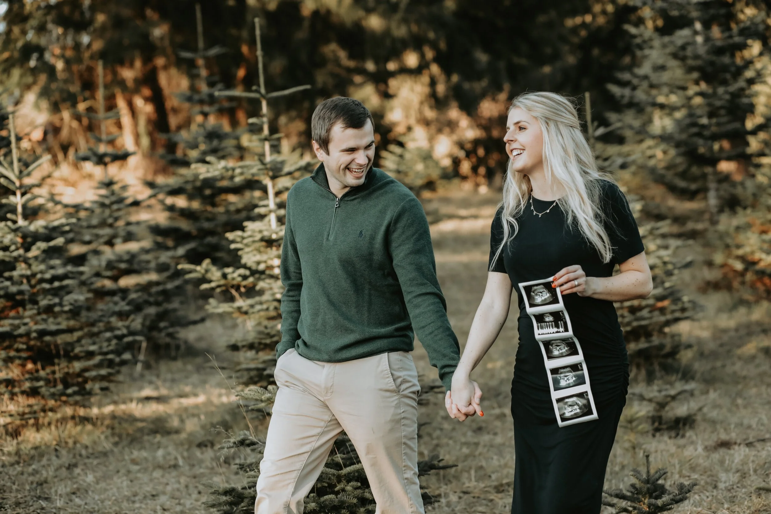 A couple walks hand in hand in a forest, the woman holding ultrasound images, both smiling at each other.