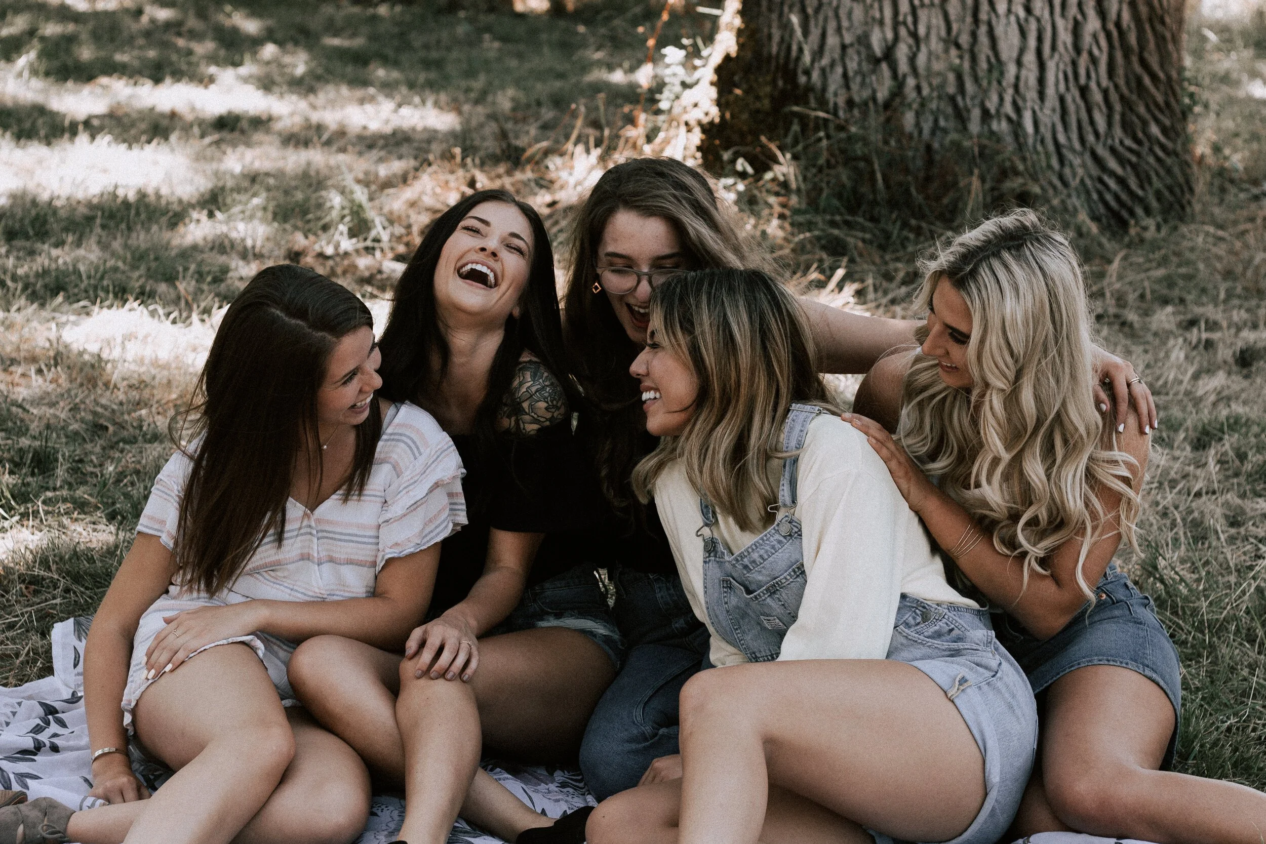 Five women sitting on a blanket under a tree, laughing and enjoying each other's company outdoors.
