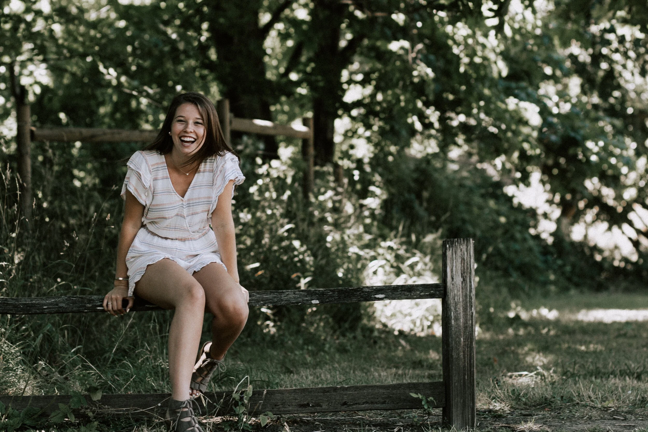 A young woman with long brown hair, wearing a light-colored, striped blouse and shorts, sitting on a wooden fence in a wooded outdoor area, laughing.