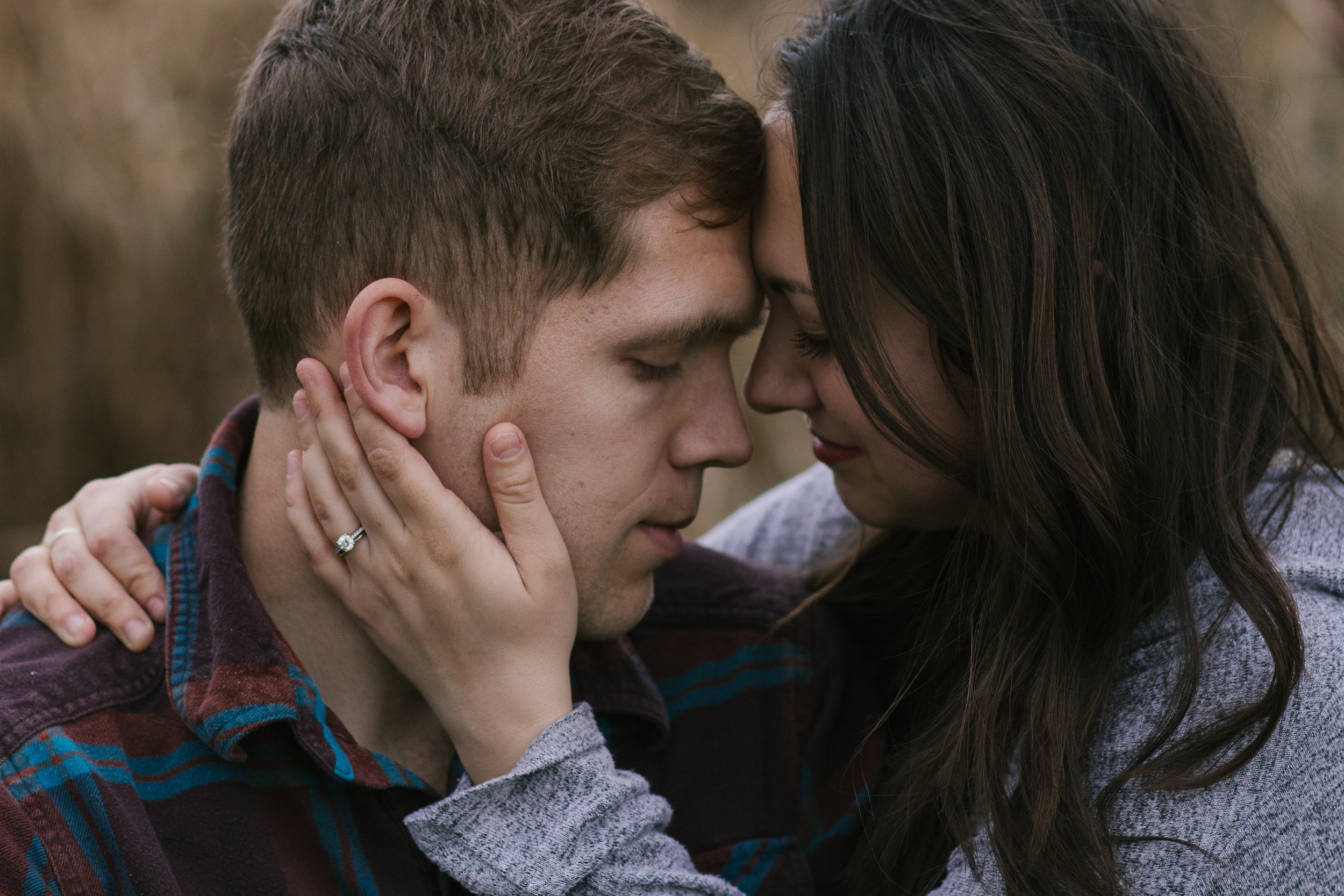 A close-up of a couple with foreheads touching, eyes closed, and hands gently holding each other's faces, in an outdoor setting.