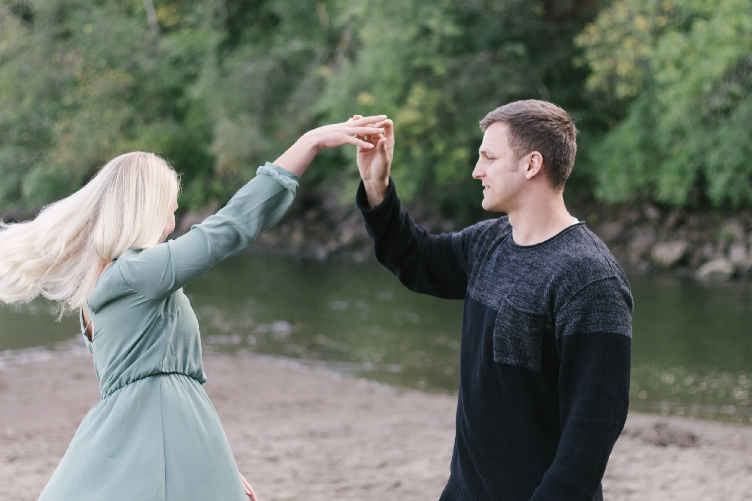 A couple dancing outdoors near a river, with the woman in a light blue dress and the man in a dark sweater, holding hands and smiling.