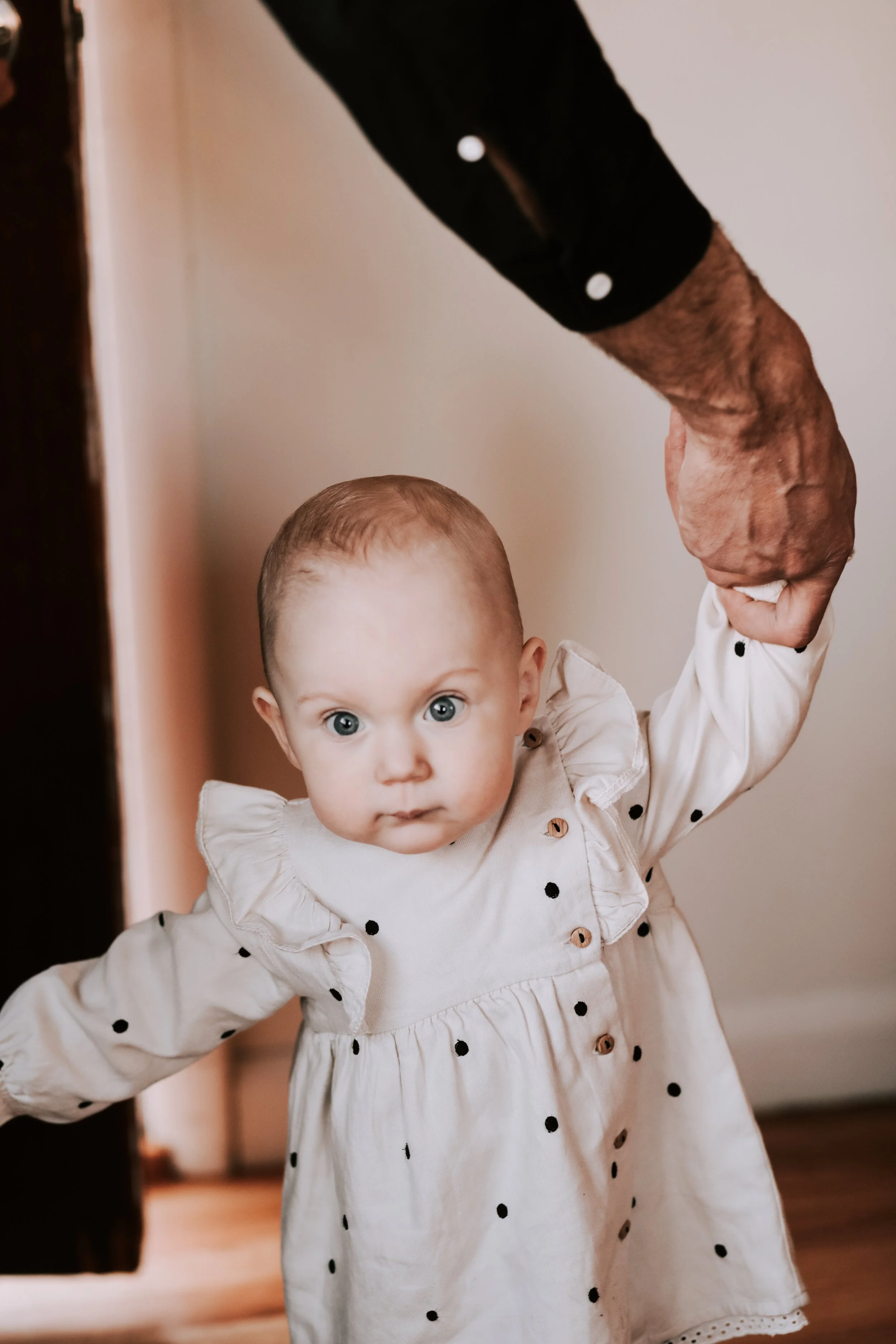 A baby girl with big blue eyes in a white dress with black polka dots is holding an adult's hand for support while standing.