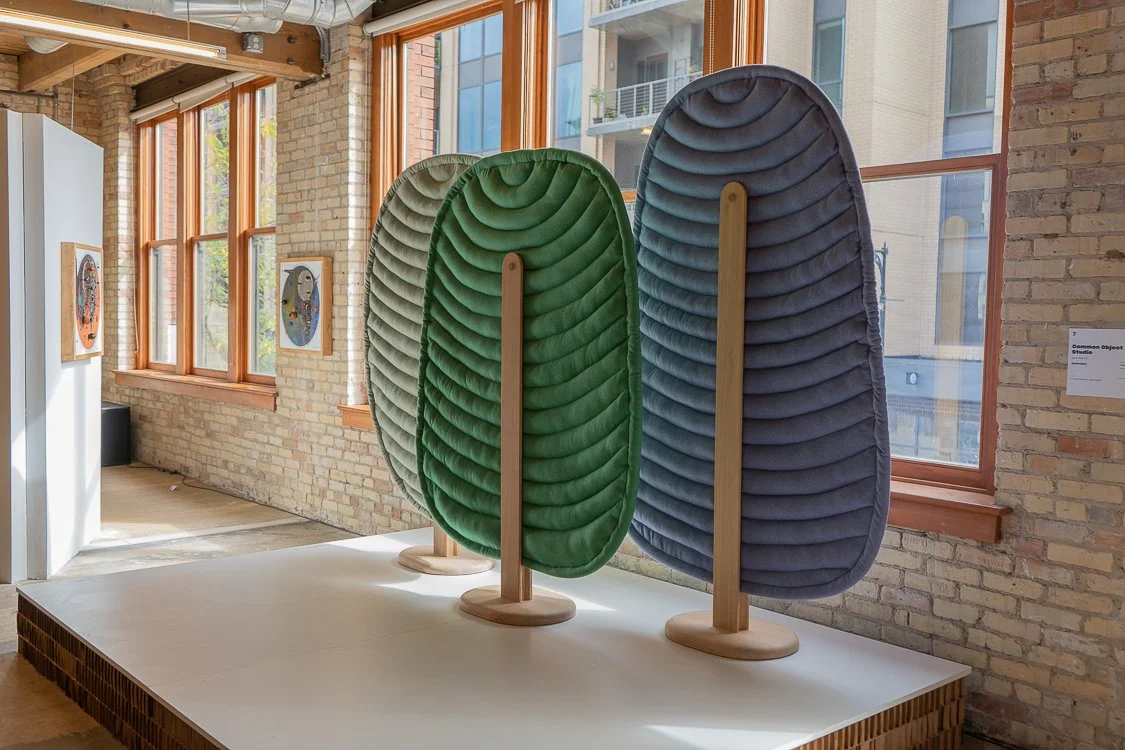 A photo of three abstract sculptures of trees. They are rounded, with layered foliage in green, gray, and darker gray. They are displayed on a white platform inside a gallery with large windows.