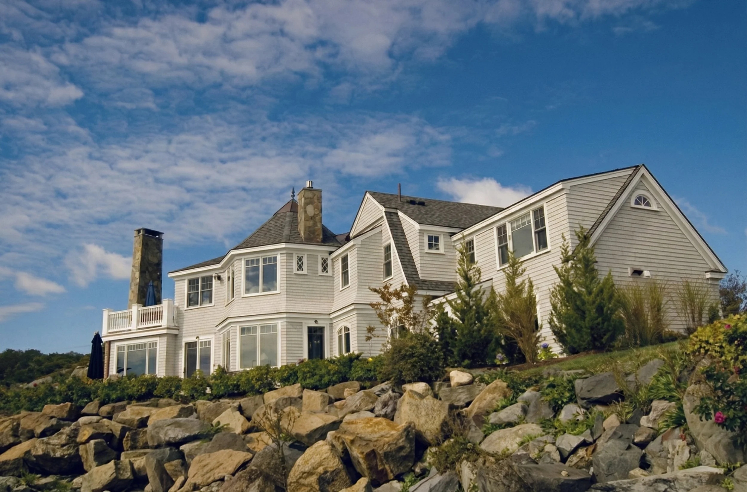 Large white house on a hill with a stone wall and garden in front, under a partly cloudy sky.