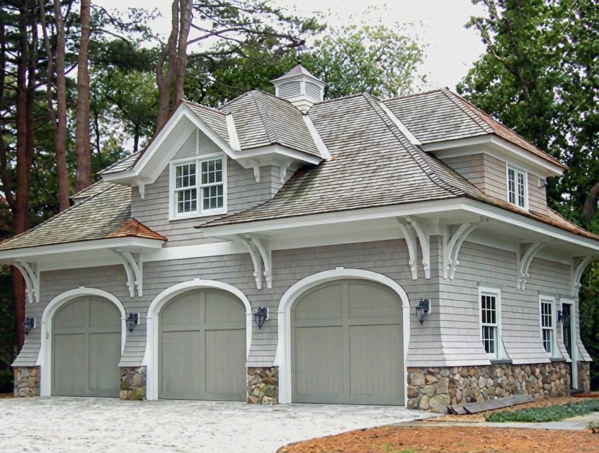 Three garage doors on a large, two-story house with light gray siding, stone foundation, and decorative white trim, surrounded by trees.
