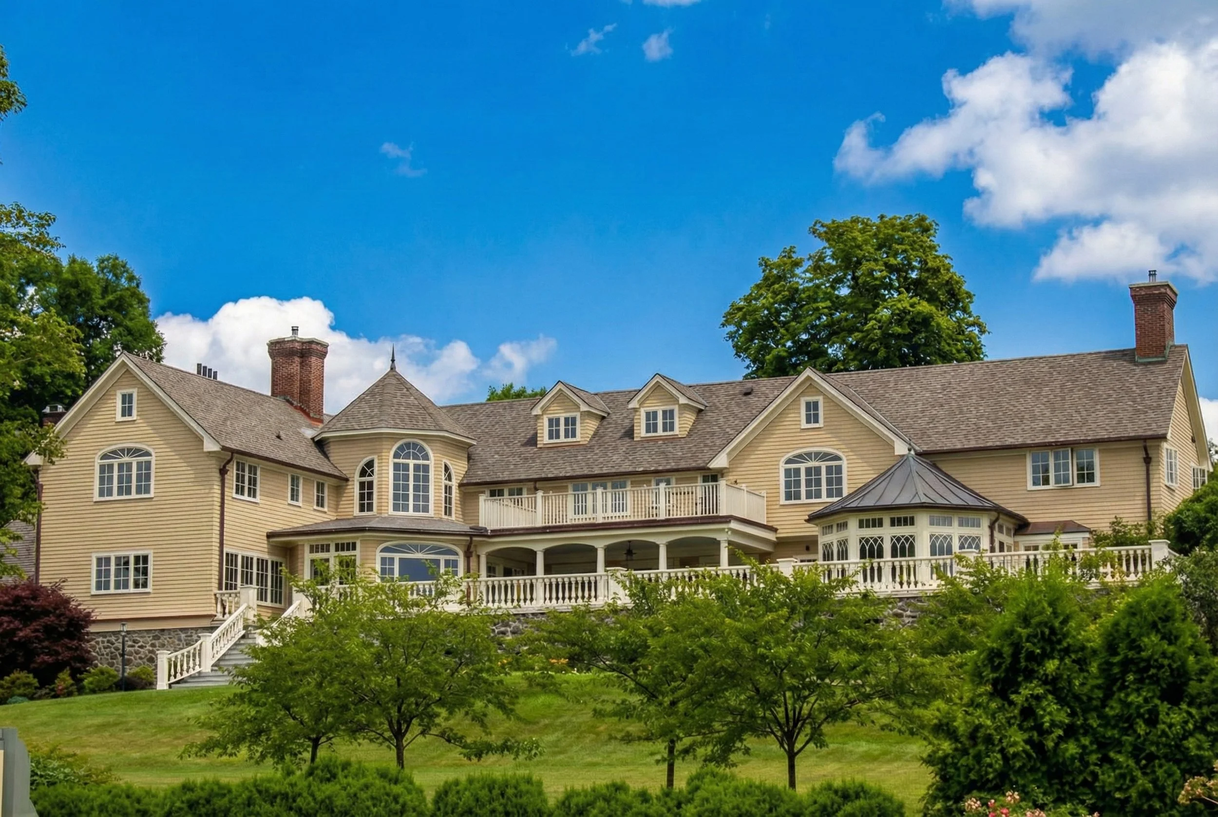 Spacious, multi-story house with beige siding, multiple gabled windows, and a wrap-around porch, surrounded by green trees and a well-maintained lawn under a bright blue sky.