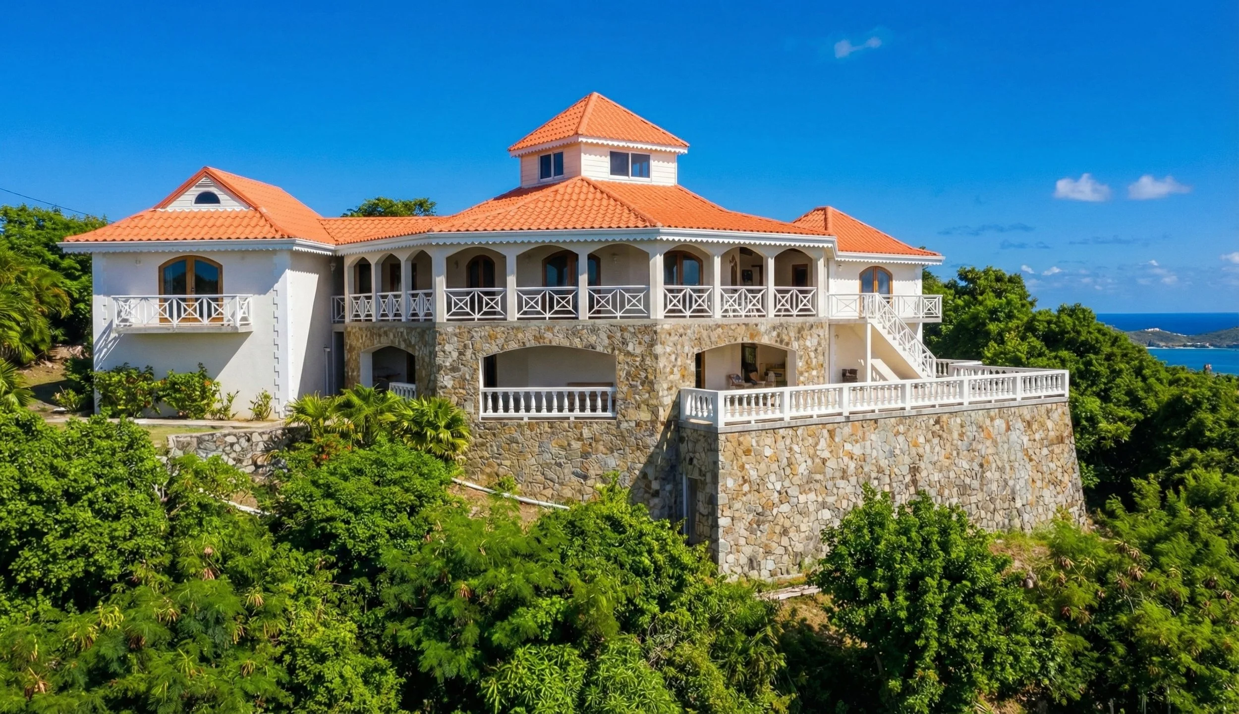 Large white house with a red tiled roof, multiple balconies, and stone foundation, surrounded by lush green trees, overlooking the ocean under a bright blue sky.