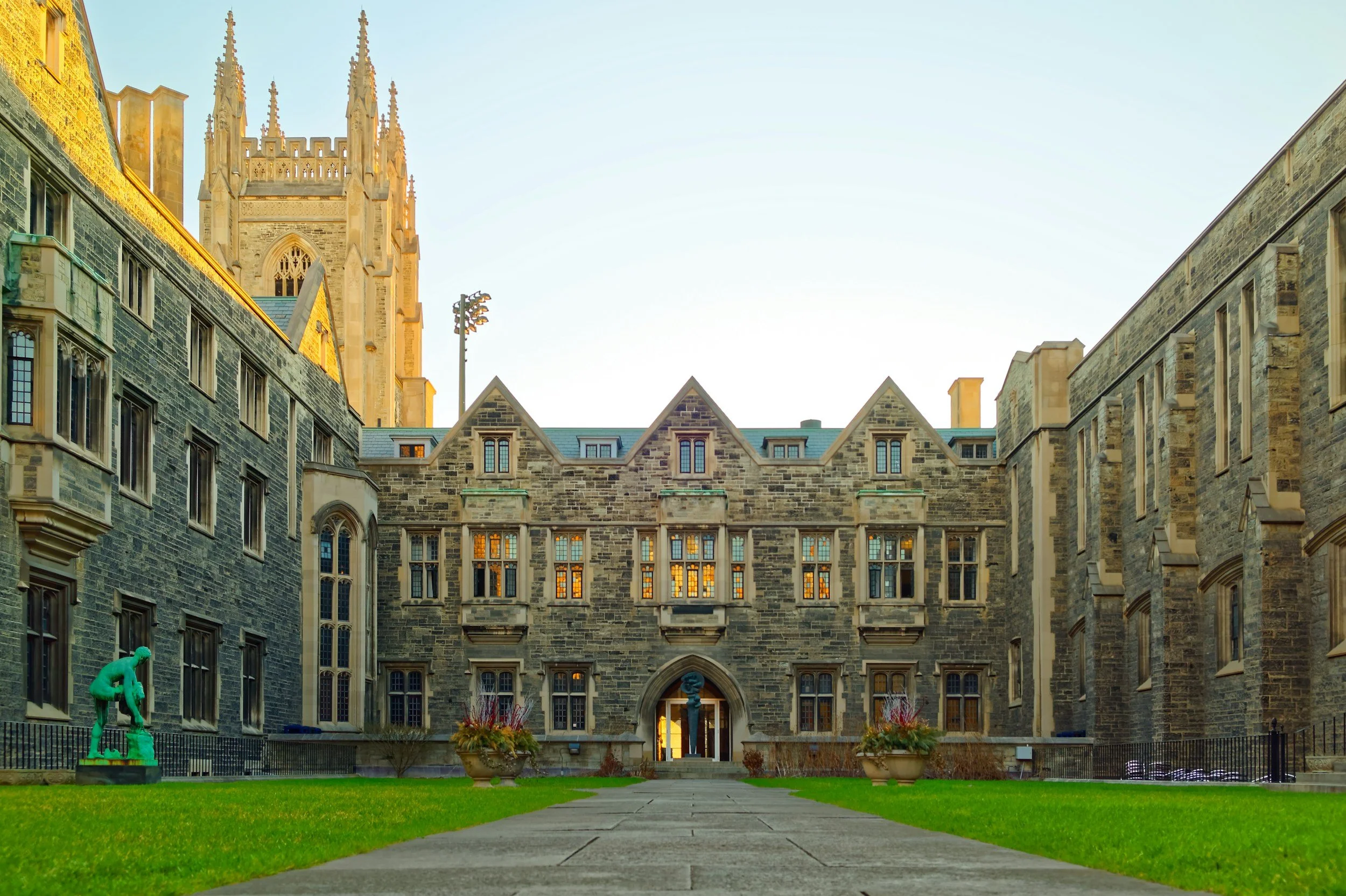 View of a historic stone building with a central entrance, gothic-style windows, and a tall tower in the background, surrounded by a well-maintained lawn with potted plants and a small statue.