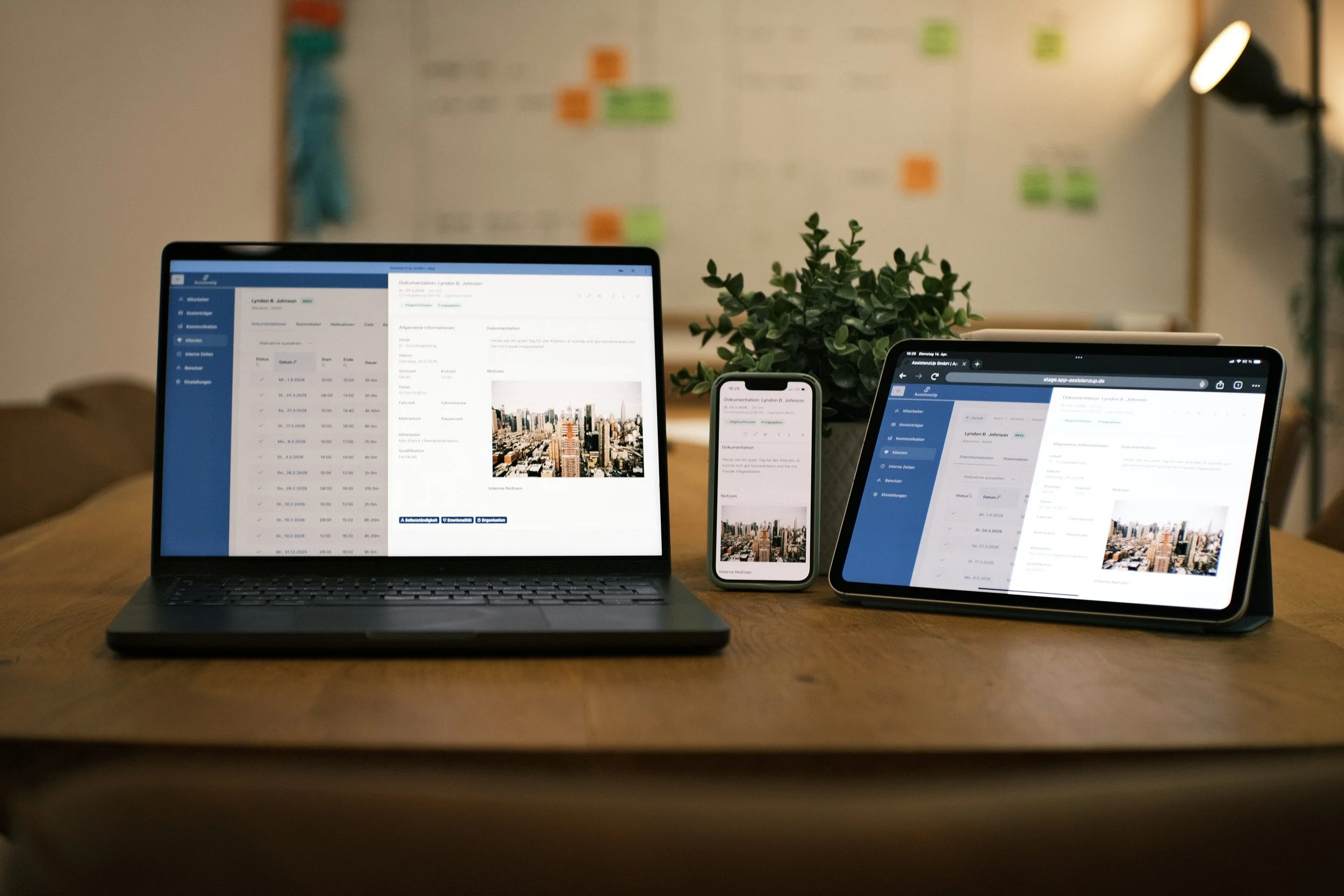 Laptop, Smartphone und Tablet auf einem Tisch, alle zeigen eine Stadtansicht und Dokumente. Im Hintergrund eine weiße Tafel mit bunten Haftnotizen. Ein grüner Pflanzenzweig steht zwischen den Geräten.