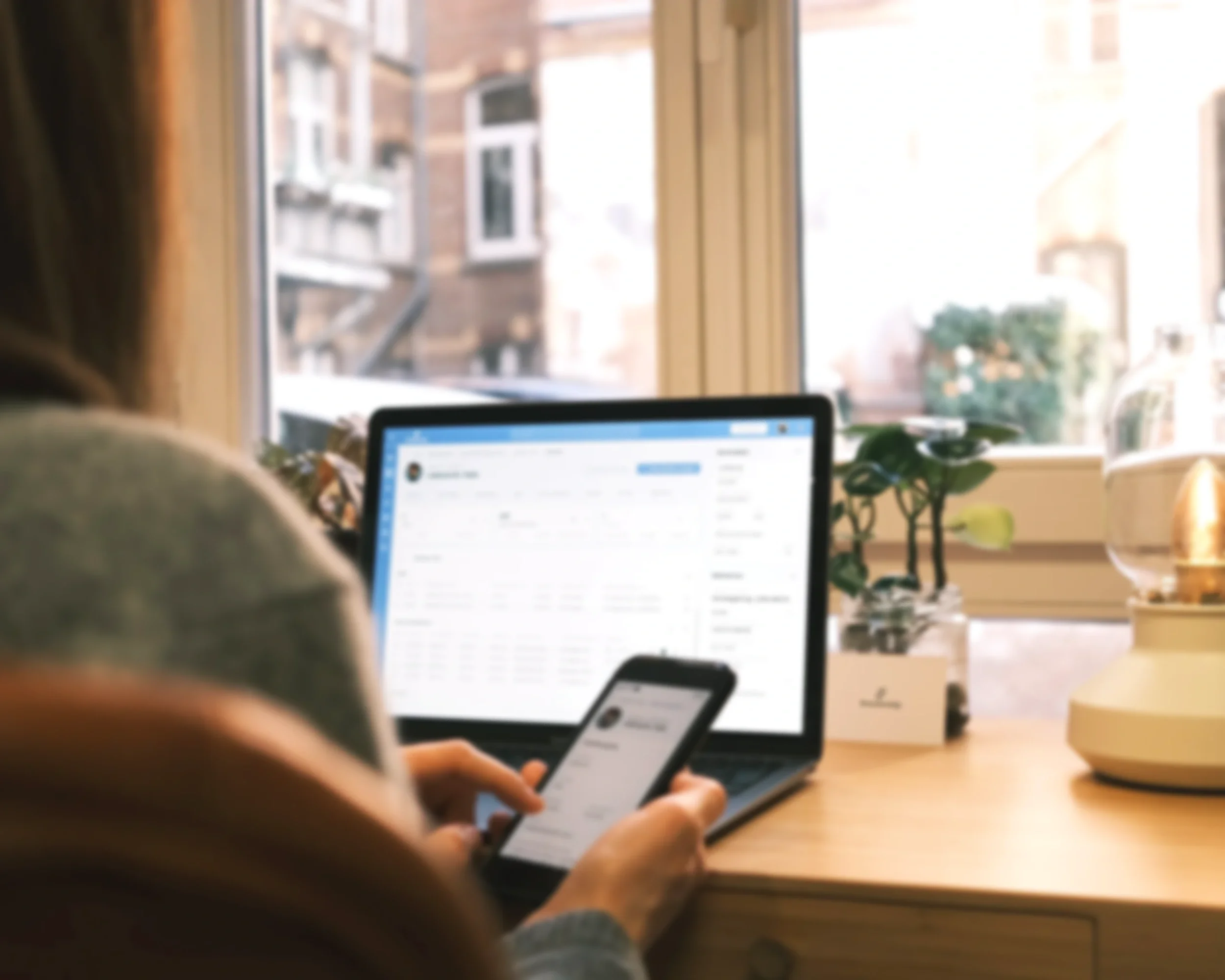 Person arbeitet an Laptop und Smartphone an einem Fenster mit Stadtblick, im Vordergrund worden die Geräte gezeigt, im Hintergrund Pflanzen und Dekoration auf dem Tisch.