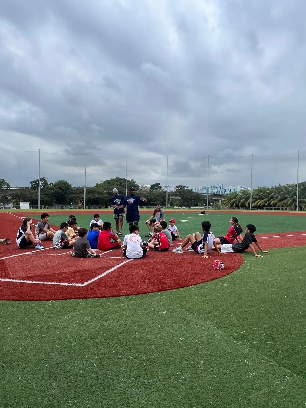 Children sitting in a circle on a baseball or softball field, listening to coaches or instructors during a practice or camp, with cloudy skies overhead.