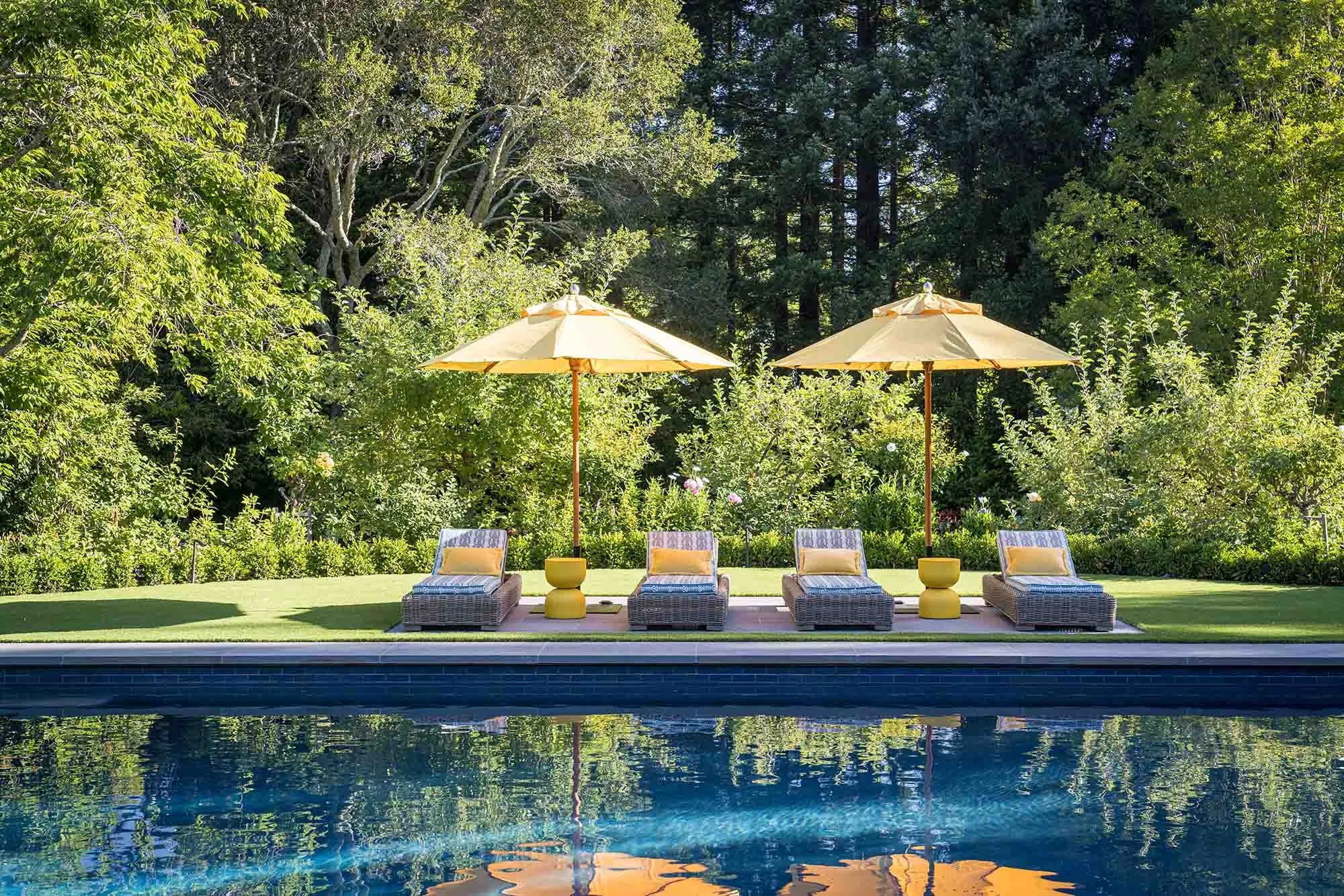 Pool deck with chaise lounges and yellow umbrellas reflecting in calm water, framed by mature trees and estate gardens