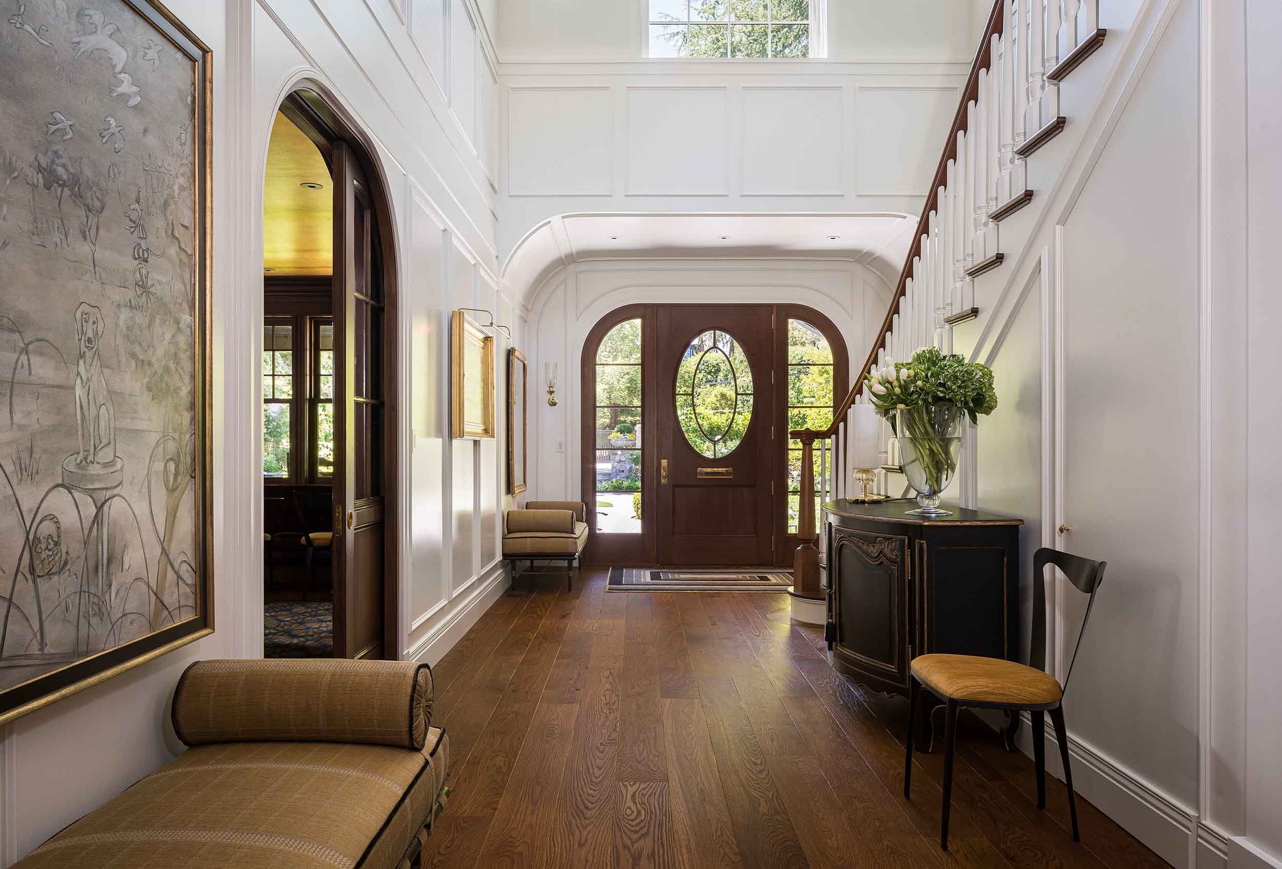 Double-height entry hall with arched front door featuring oval leaded glass window, wide plank hardwood floors, and wainscoted walls. 