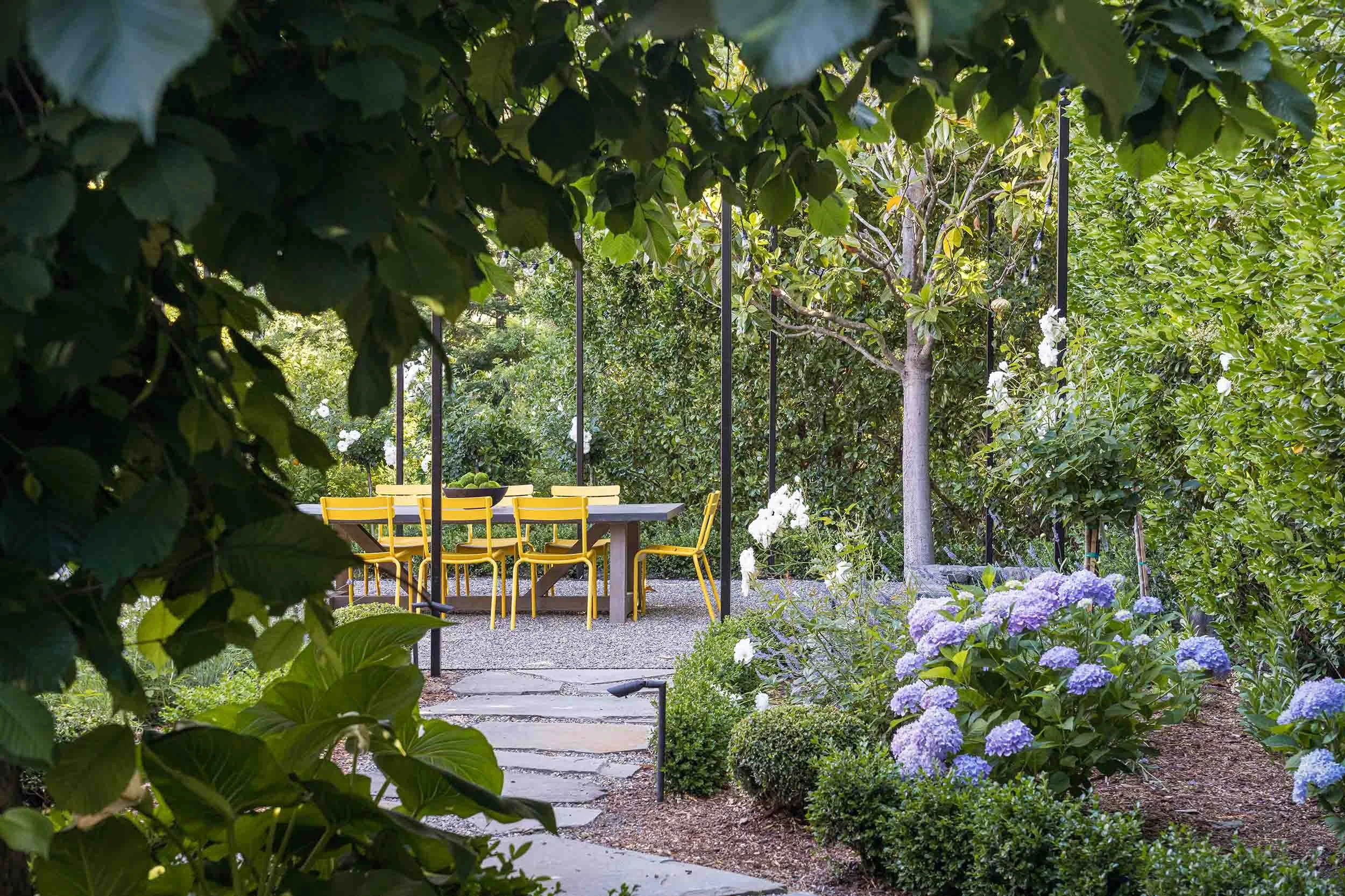Garden path view of outdoor dining area framed by hydrangeas, magnolia trees, and string lights 