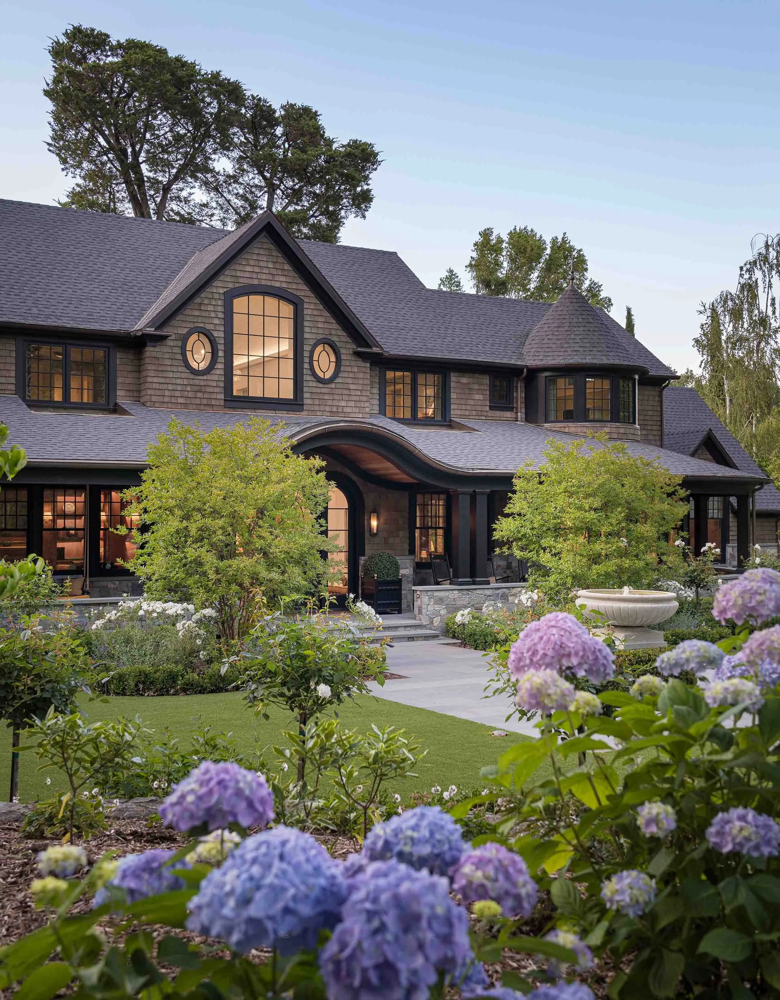 Norman Charles Construction shingle style custom home twilight exterior with hydrangeas, arched cedar entry, and blue evening sky.
