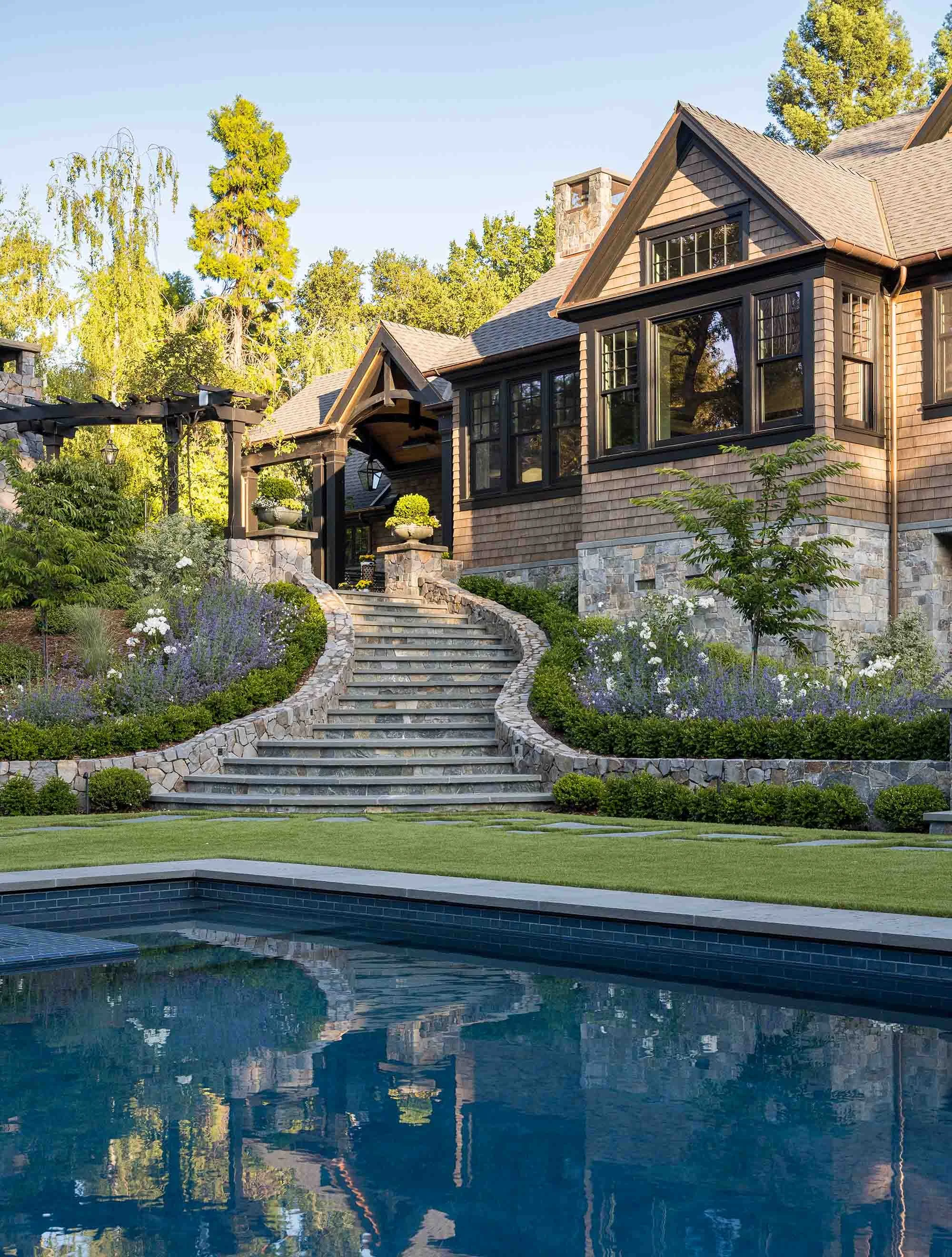 Custom swimming pool with terraced stone steps, curved landscaping, and Norman Charles Construction shingle style home
with pergola in background.