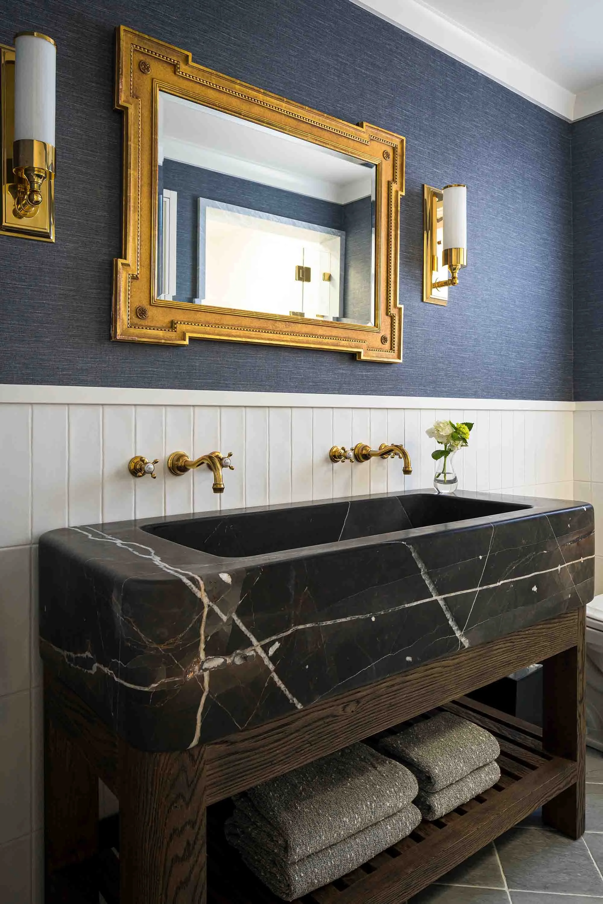 Powder room with black marble trough sink on wood vanity, gold framed mirror, brass wall-mount faucets, and navy grasscloth walls 