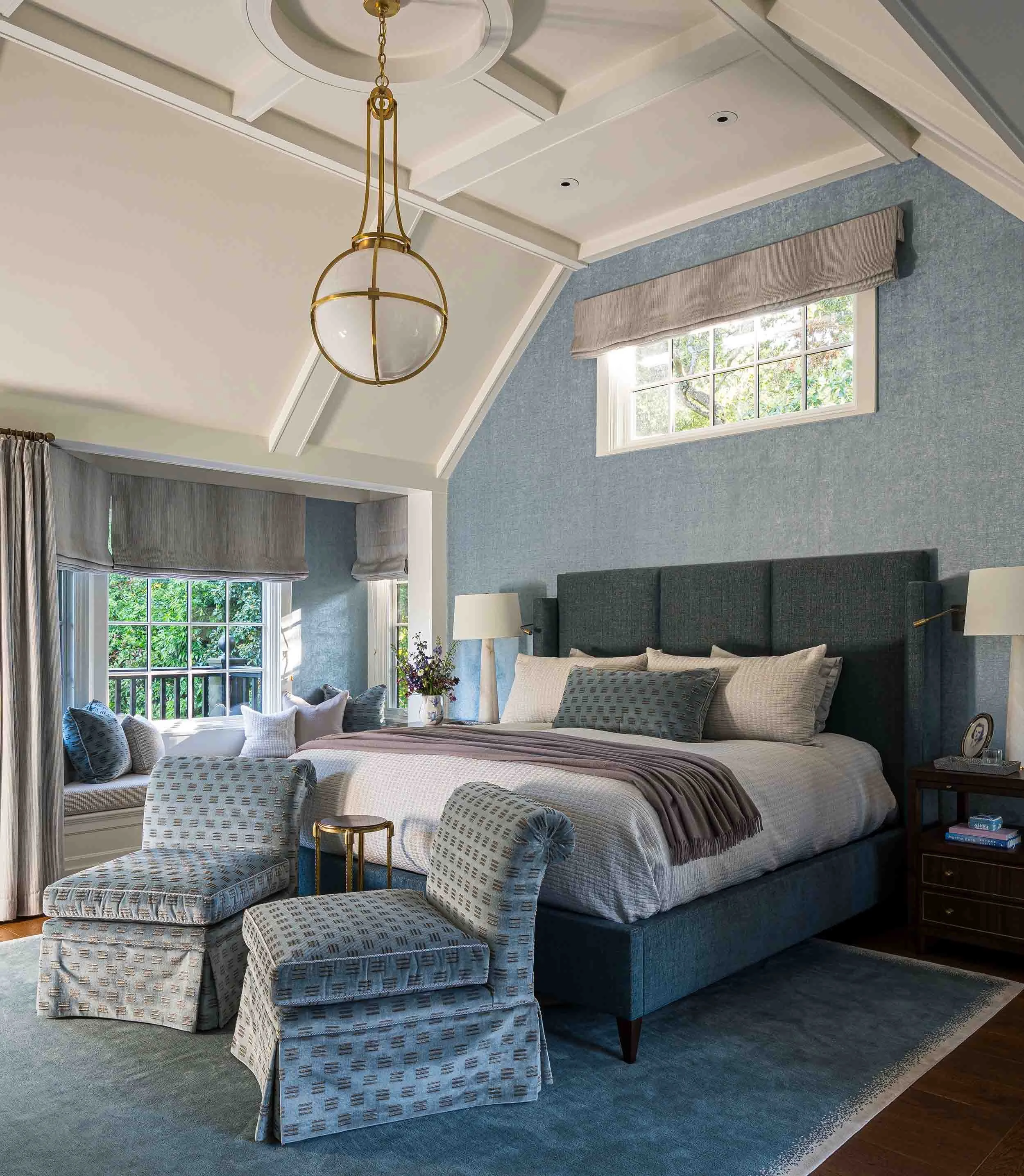 Primary bedroom with ornate coffered vaulted ceiling, blue grasscloth walls, upholstered headboard, and brass globe pendant. 
