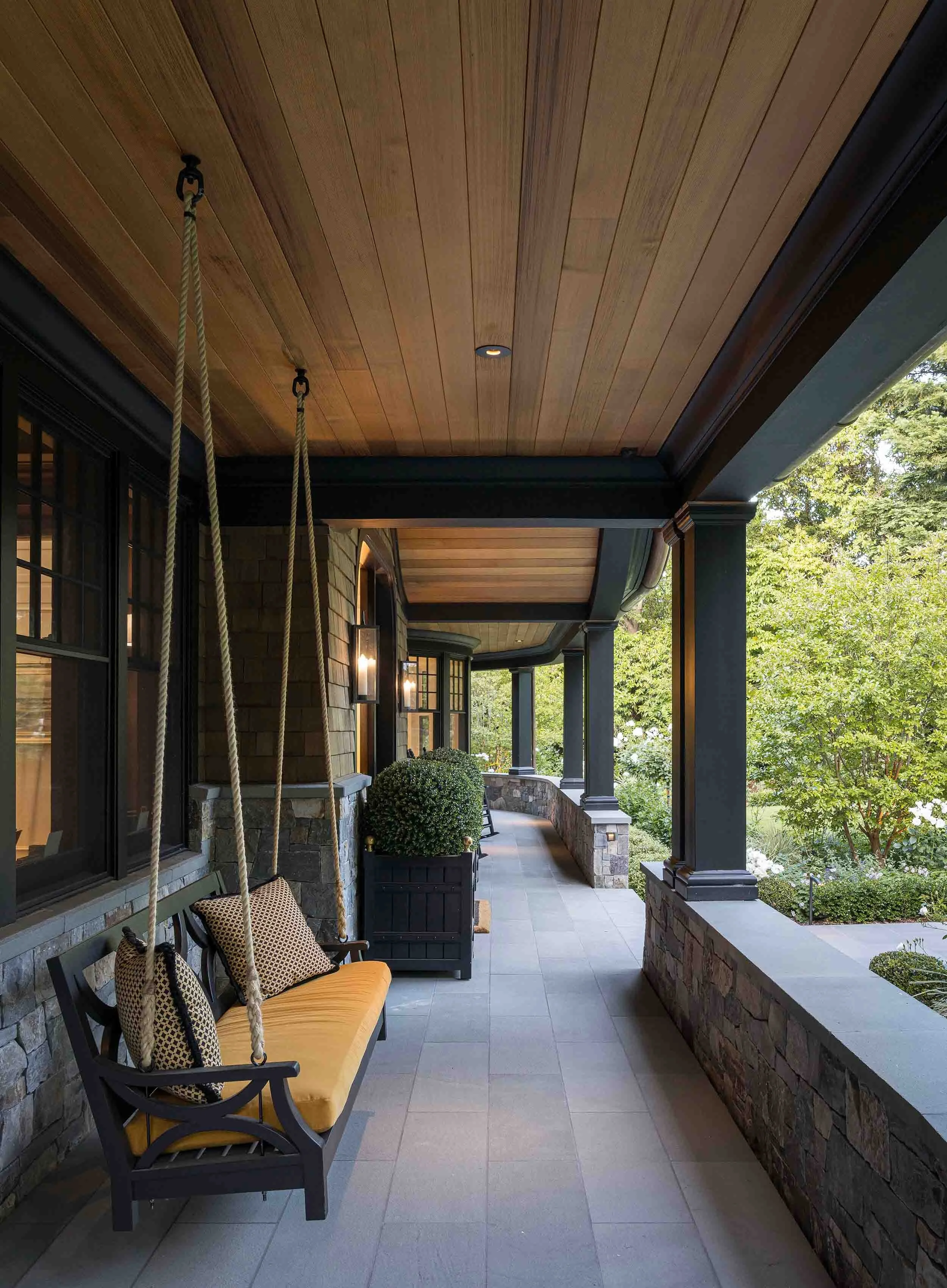 Covered side porch with cedar tongue-and-groove ceiling, rope porch swing with cushions, and stone half-wall