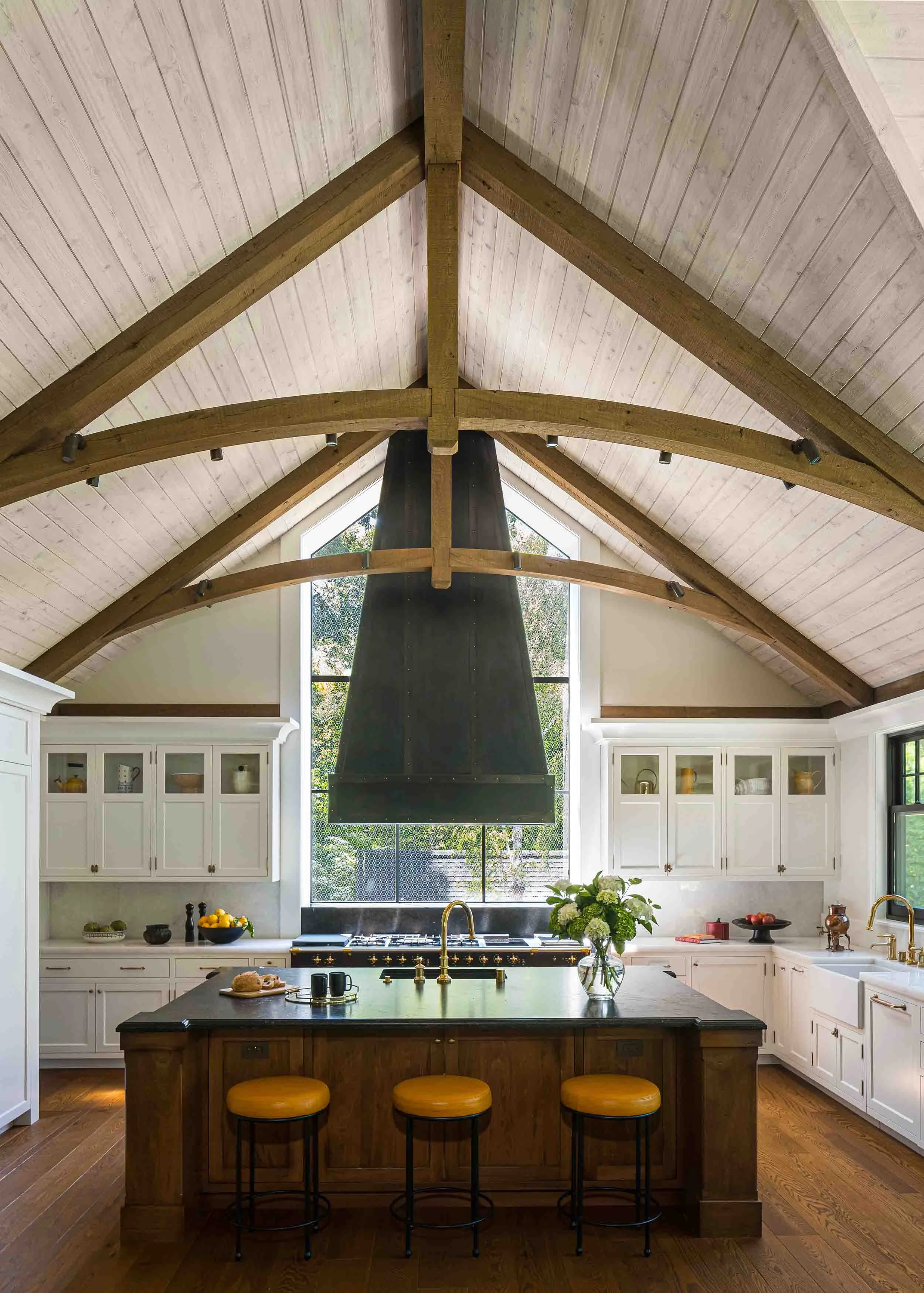 Custom kitchen with dramatic vaulted ceiling, exposed timber trusses, walnut island, leather bar stools, and oversized black range hood in Norman Charles Construction home.