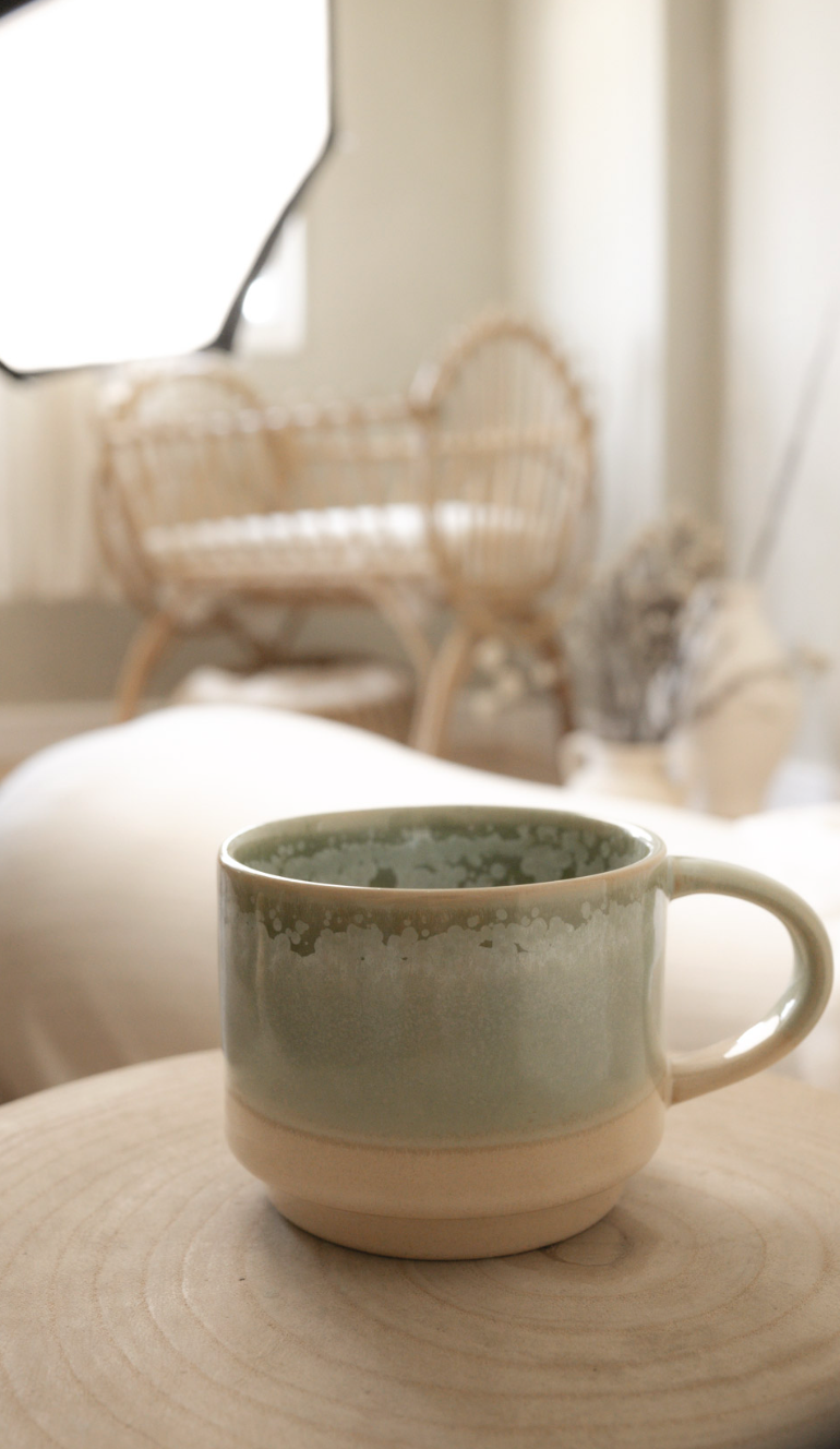 A ceramic mug with a greenish glaze on top and a cream-colored base, placed on a wooden surface in a cozy room with wicker furniture and neutral decor.