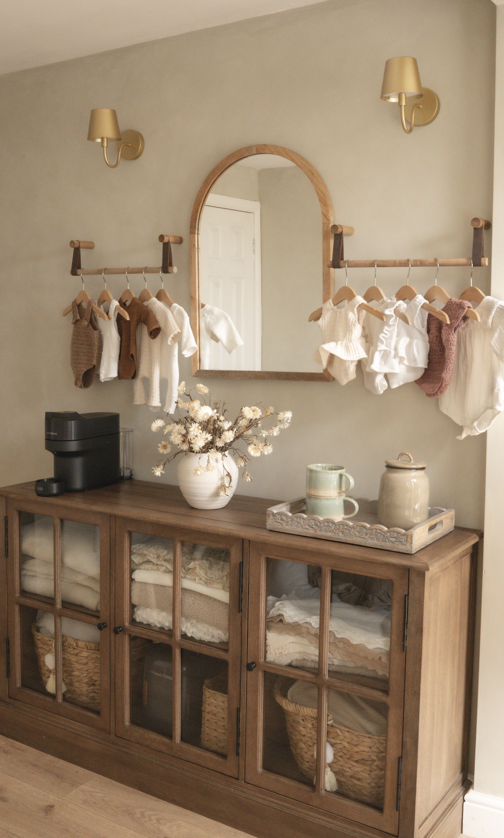 Wooden cabinet displaying newborn wraps and baskets beneath a mirror, with a coffee station and neutral baby clothing hanging on either side in Little River Photography's studio.