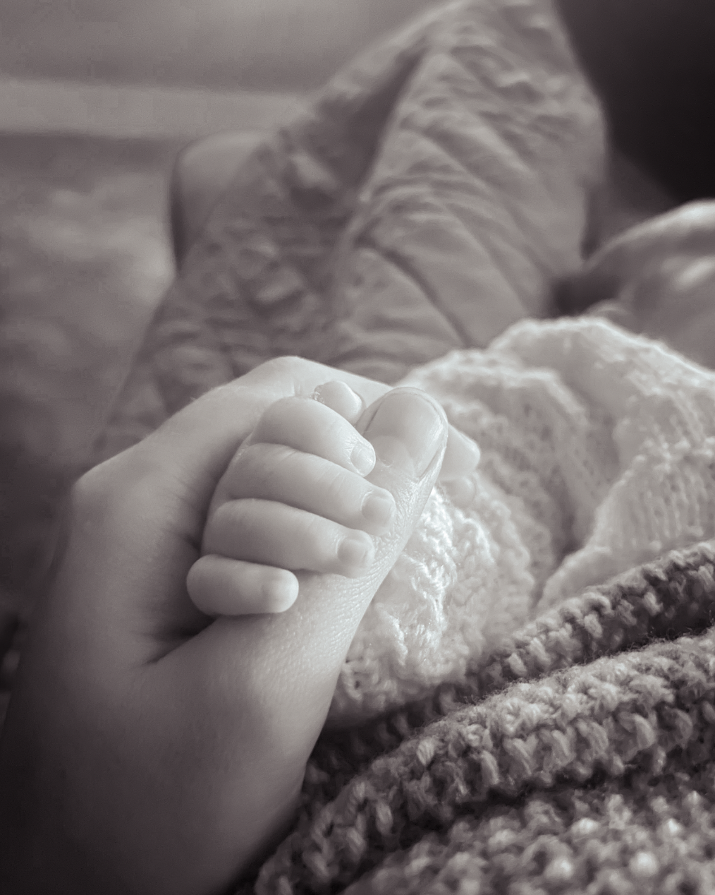 A black and white photo of a child's hand holding an adult's finger, with blankets in the background.