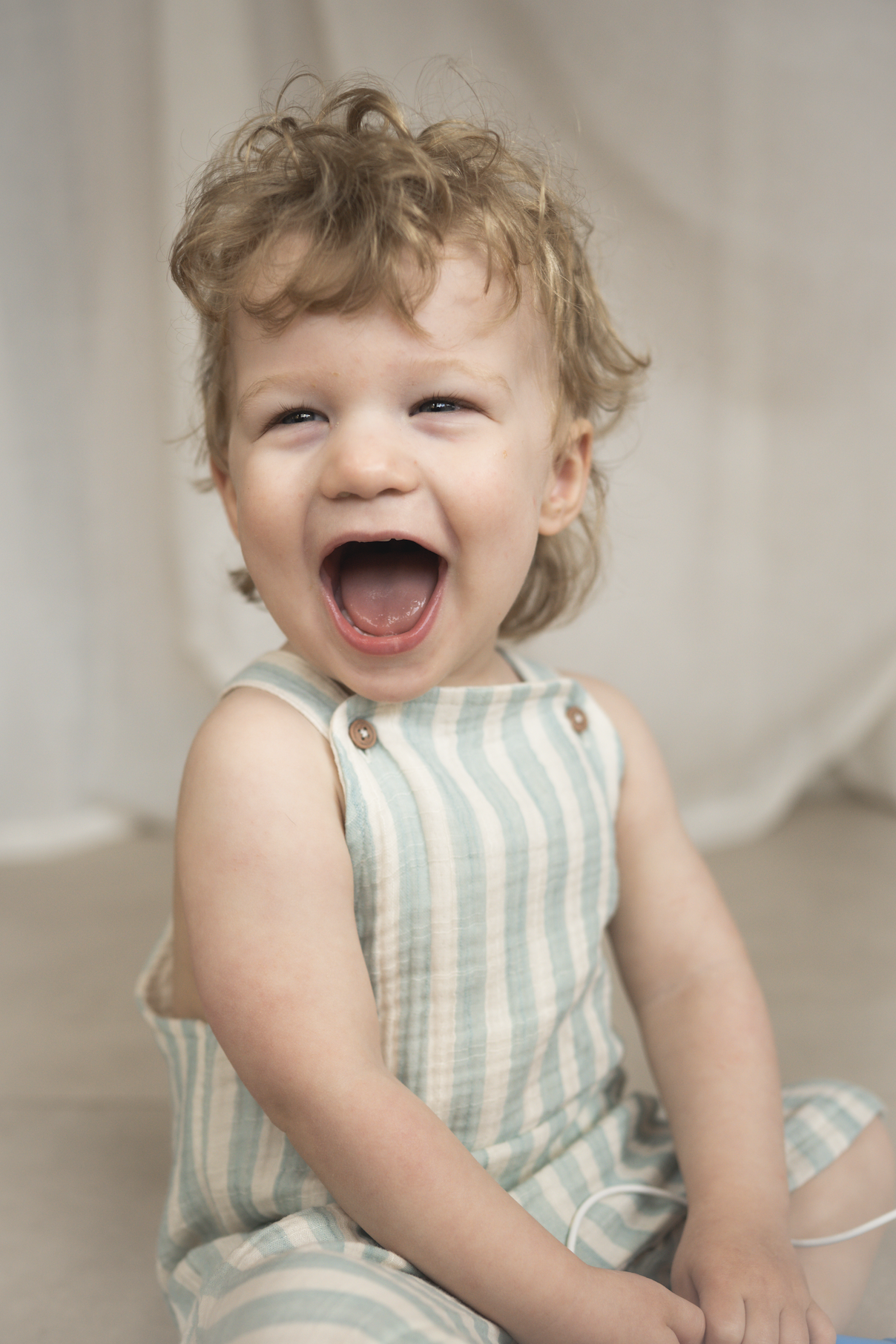 A young child with curly hair wearing a striped sleeveless outfit, laughing open-mouthed, sitting on the floor indoors.