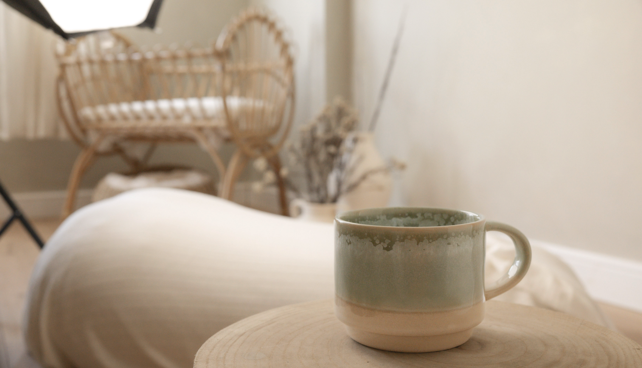 A beige ceramic mug with a speckled glaze  with a background featuring a bamboo cot chair, dried flowers, and a wall.