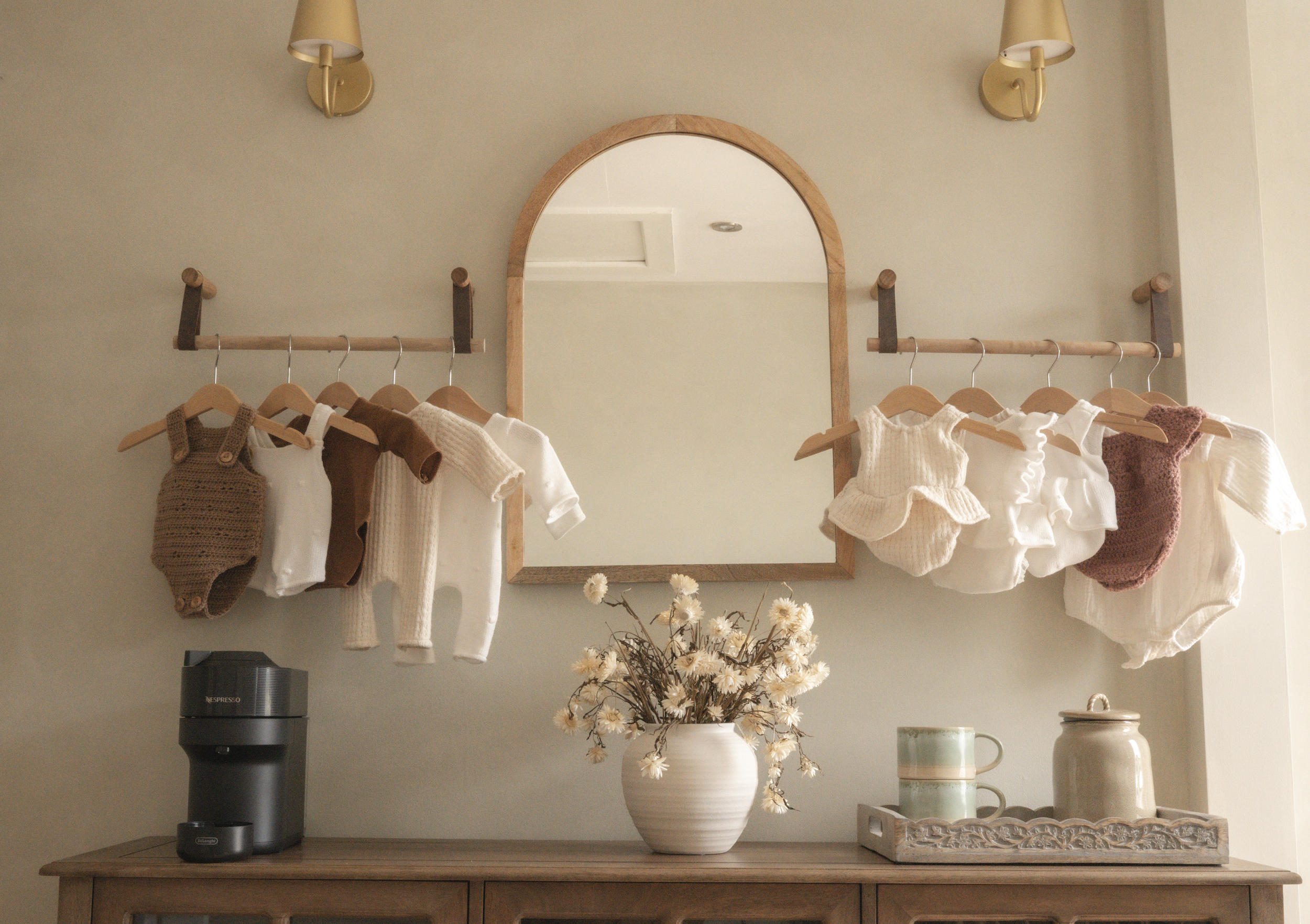 Wooden cabinet with folded newborn wraps and baskets, topped with a coffee station, mirror and hanging baby clothing in soft neutral tones at Little River Photography.