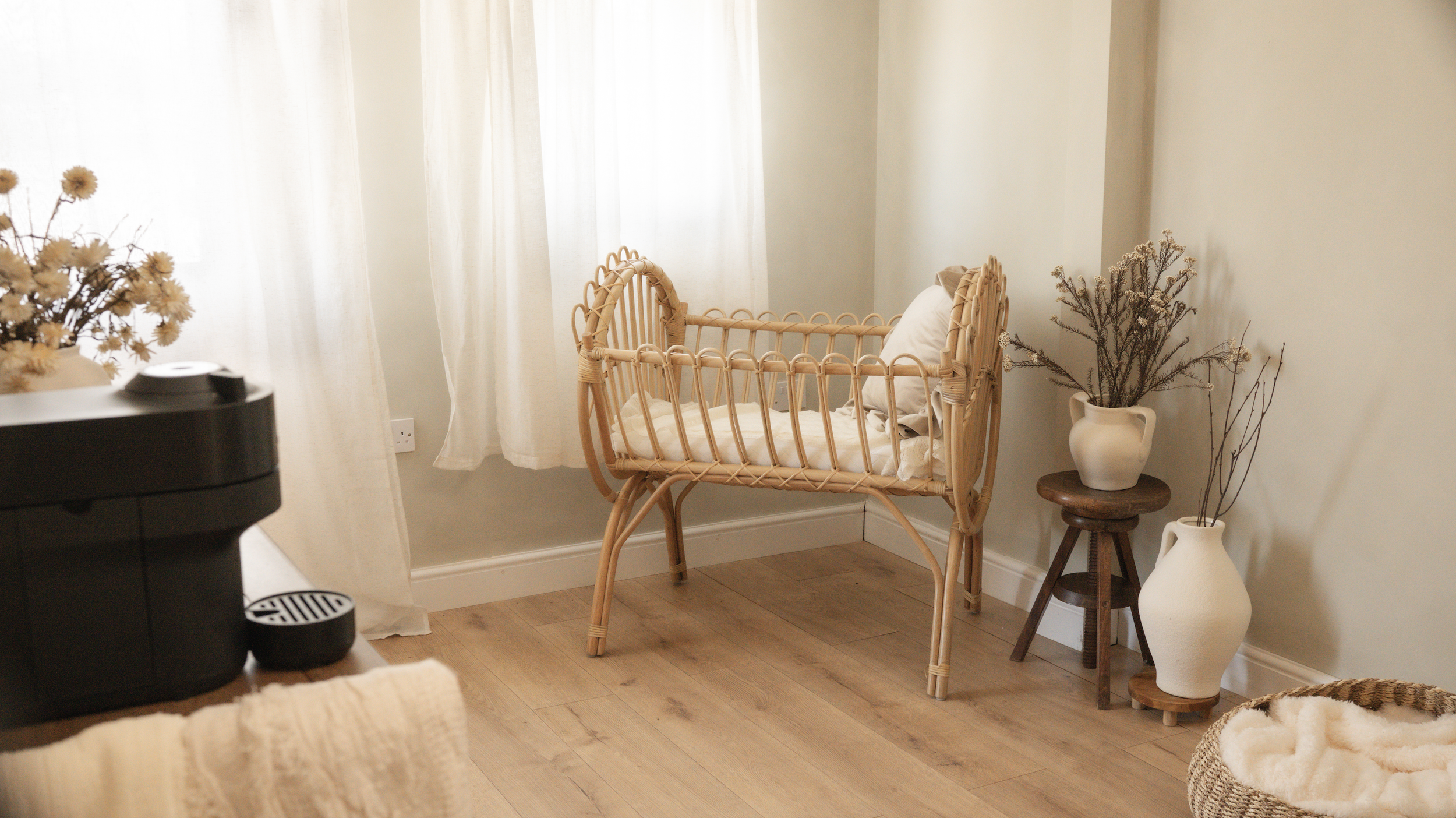 A cozy room corner with a rattan crib, white curtains, and decorative vases with dried flowers.