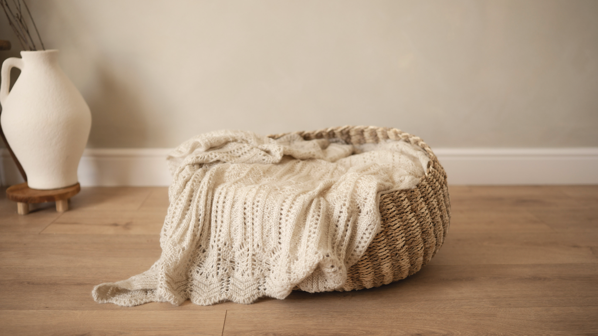 A woven basket with a knitted cream-colored blanket draped over it, positioned on a wooden floor against a plain wall. To the left, there is a white ceramic vase on a small wooden stand, with some dried branches inside.