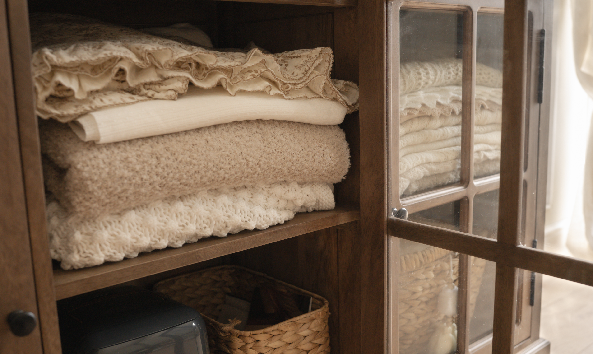 Wooden cabinet with glass door containing neatly folded beige and cream wraps and blankets.