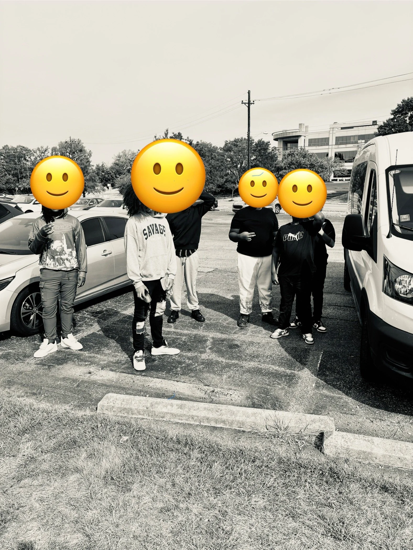 Group of seven children standing in a parking lot, with their faces covered by smiley face emojis. The children are dressed casually and are standing near parked cars and a white van.