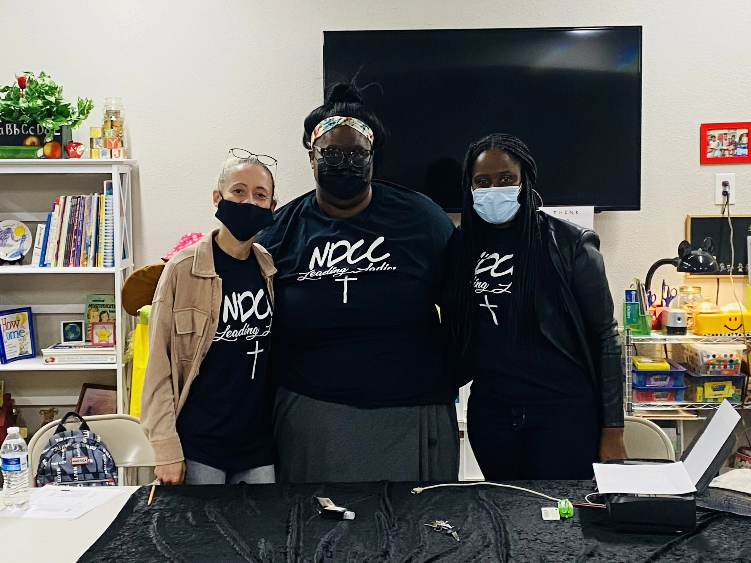 Three women standing together in a classroom or office, all wearing black t-shirts with white lettering and crosses, and face masks. There are bookshelves, a television, and various supplies in the background.