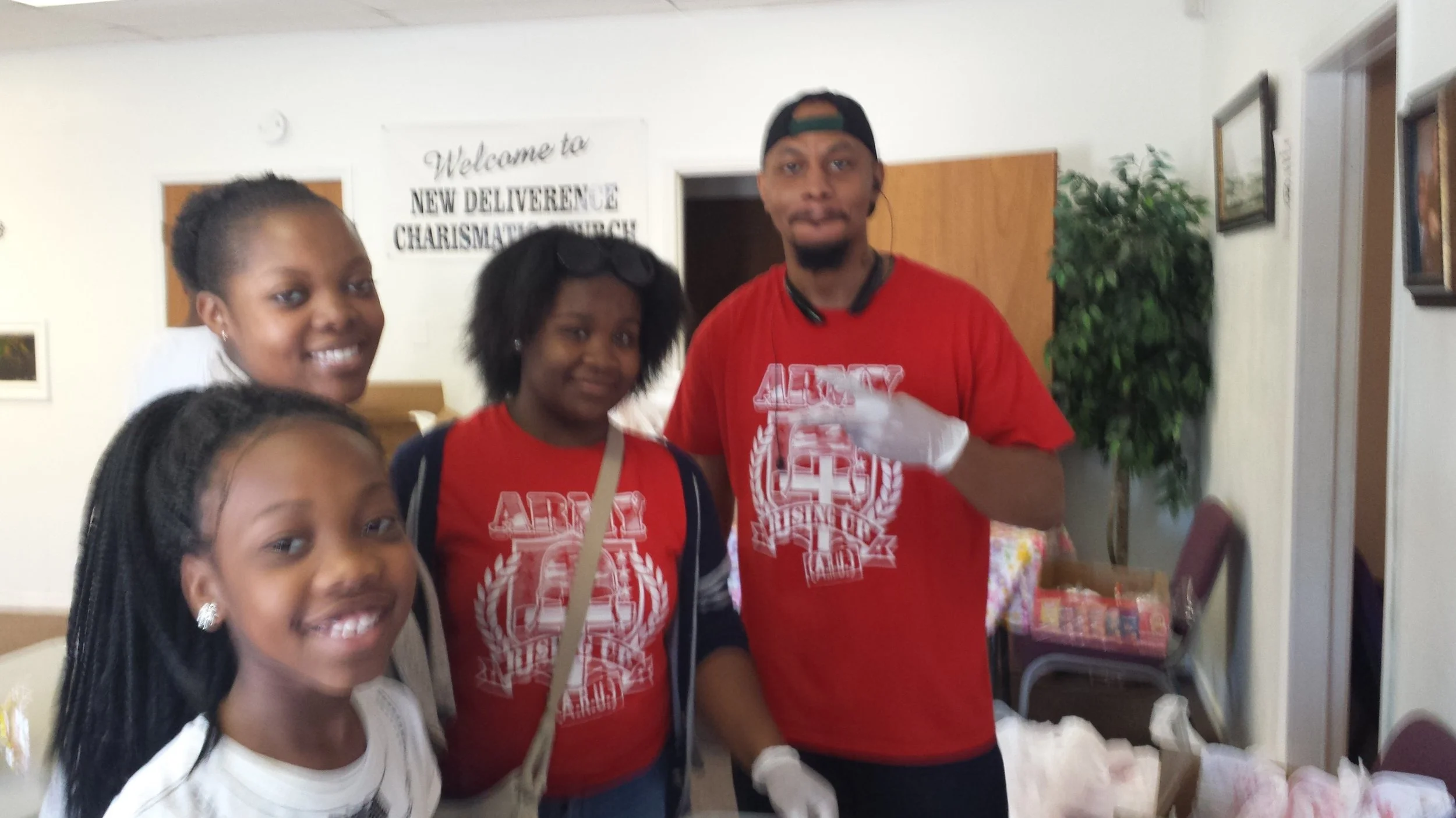 Group of four people, three women and one man, standing together indoors. They are smiling and pose for a photo. Behind them, a sign reads 'Welcome to New Deliverance Charismatic Church.' Some furniture and decorations are visible in the background, along with items on a table in the foreground.