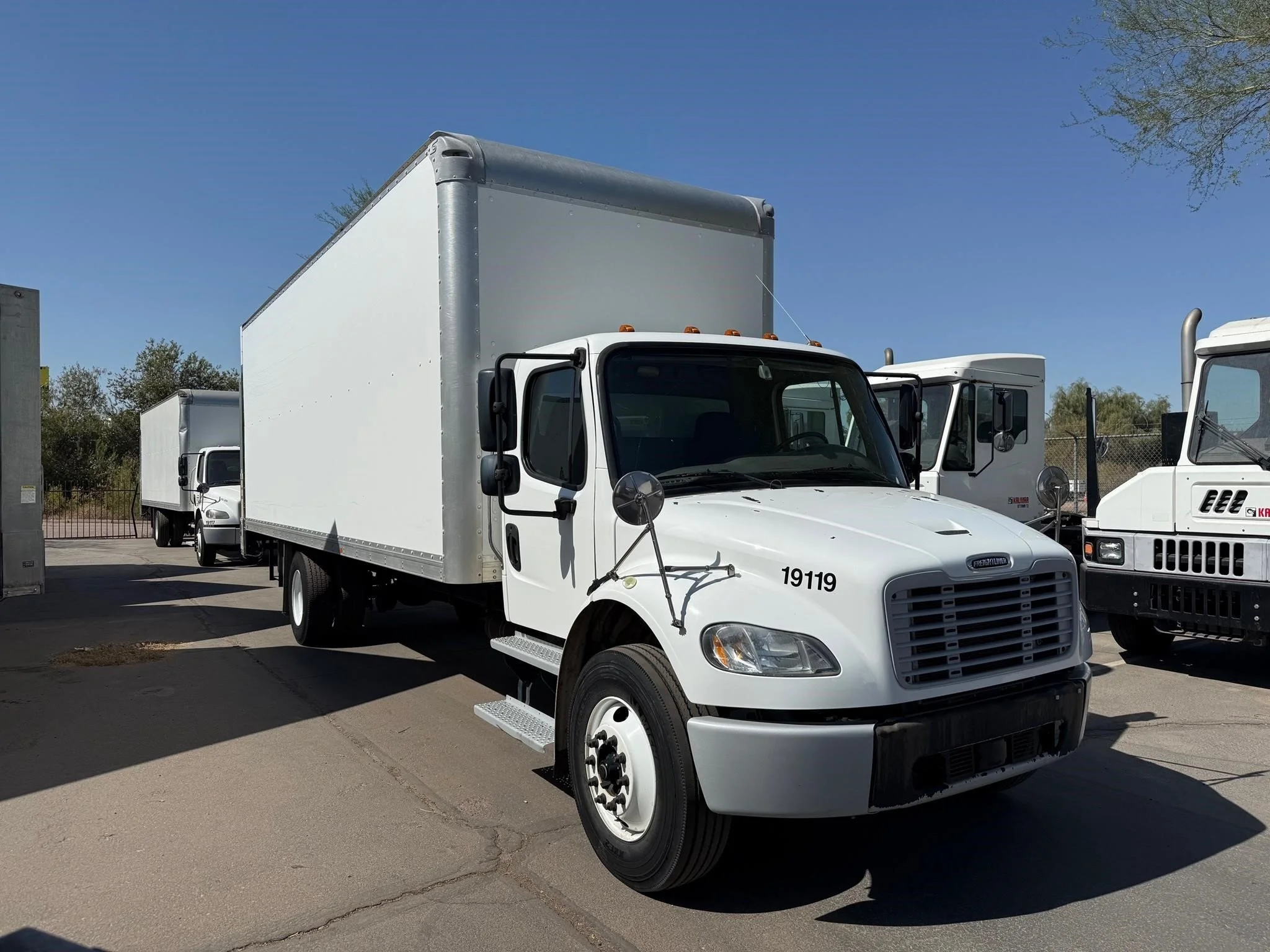 White box truck parked on a lot with two similar trucks in the background under a clear blue sky.