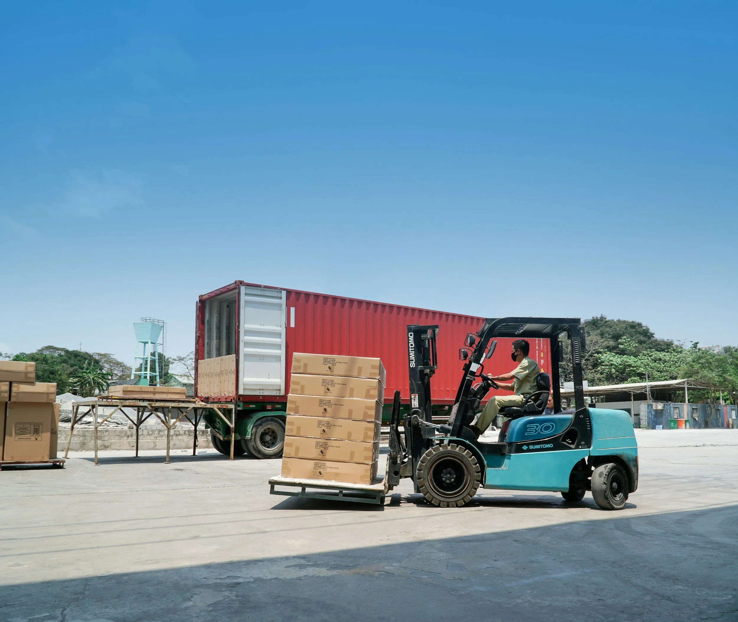 A person operating a blue forklift transporting cardboard boxes in an outdoor warehouse area, with a red truck parked nearby and several crates and boxes scattered around.