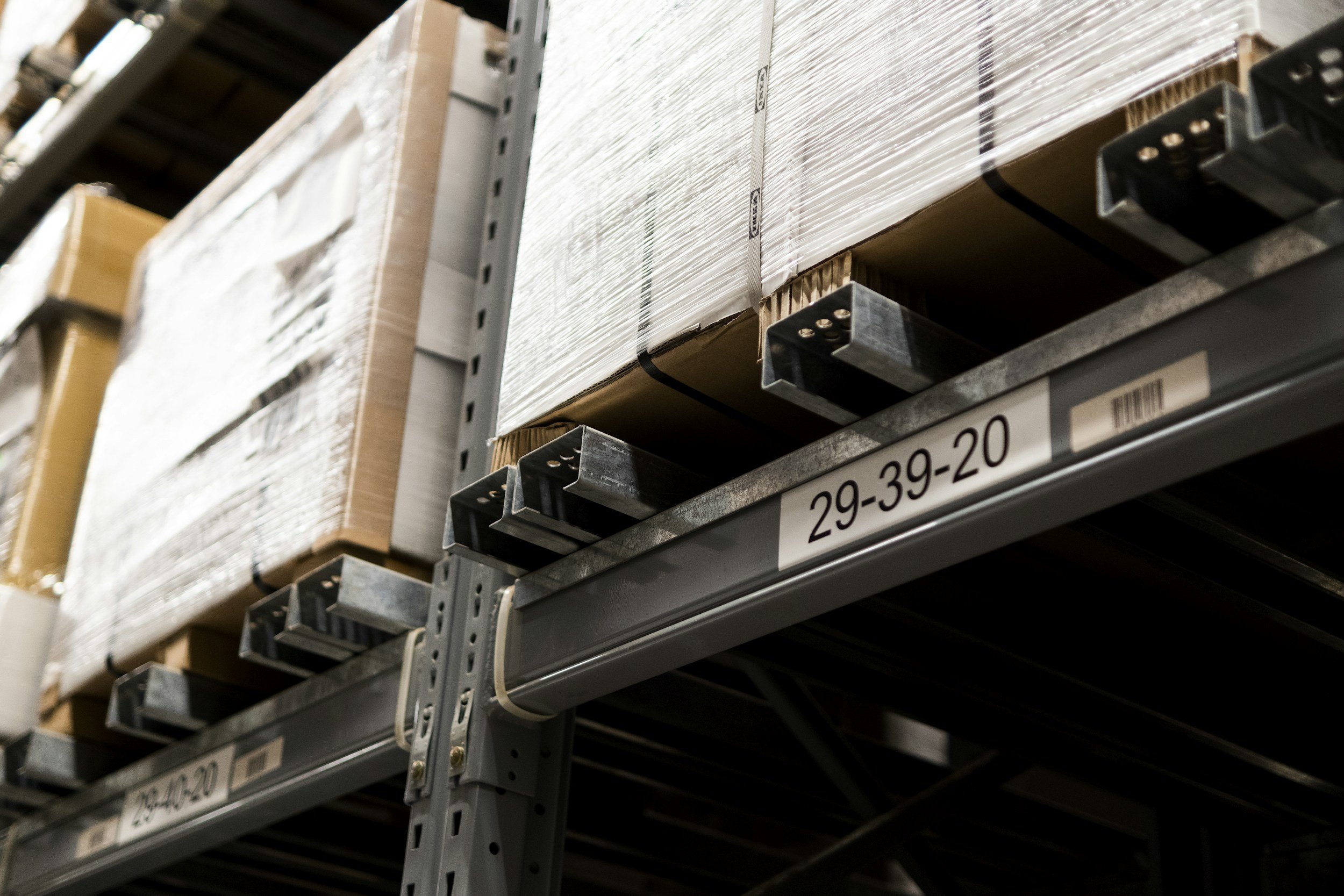 Close-up of warehouse metal shelving filled with large cardboard boxes wrapped in plastic, with labeled metal shelf supports showing the numbers 29-39-20 and 29-39-20.