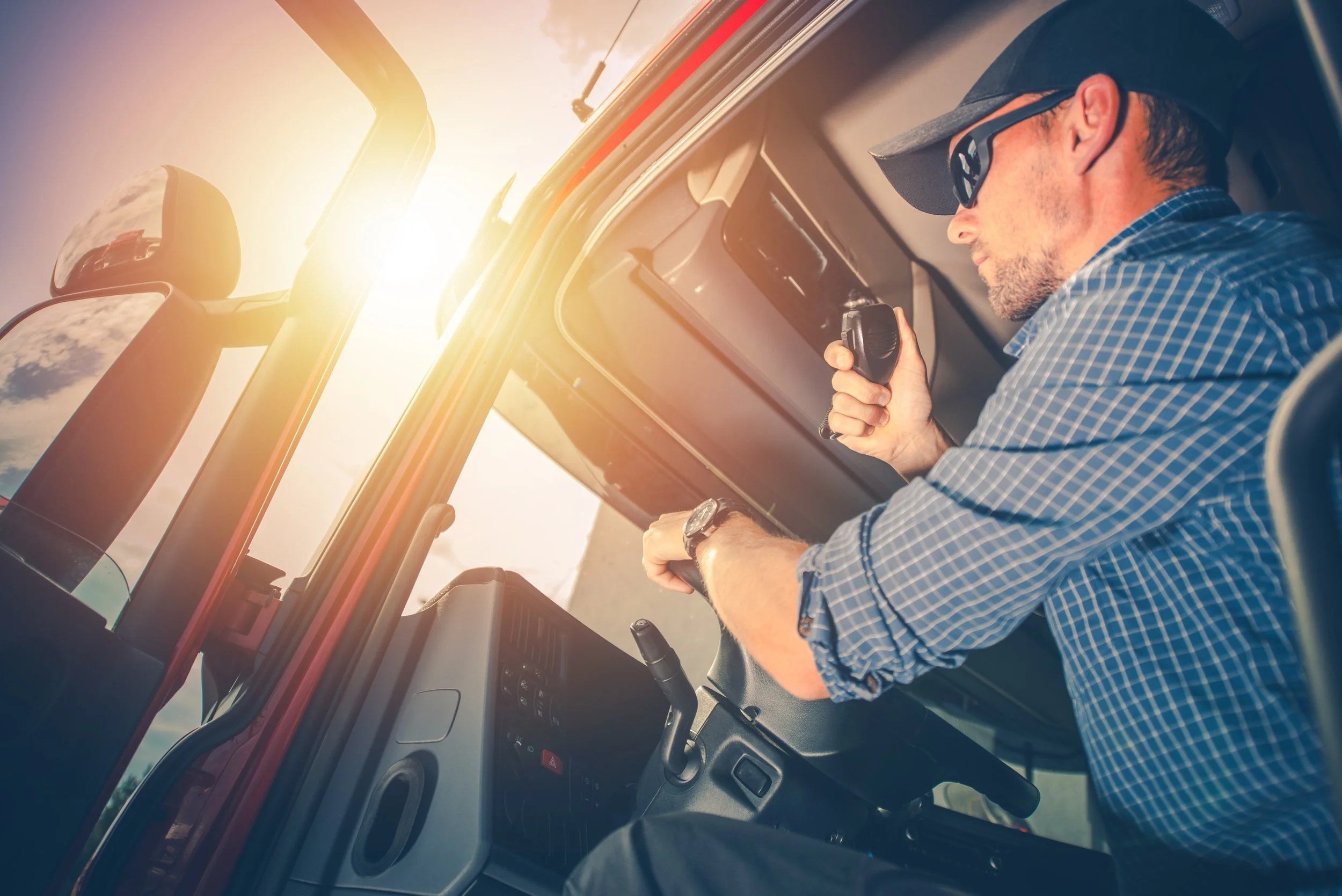 A man sitting in the driver's seat of a truck, looking at a handheld radio device with sunlight shining in through the windshield.