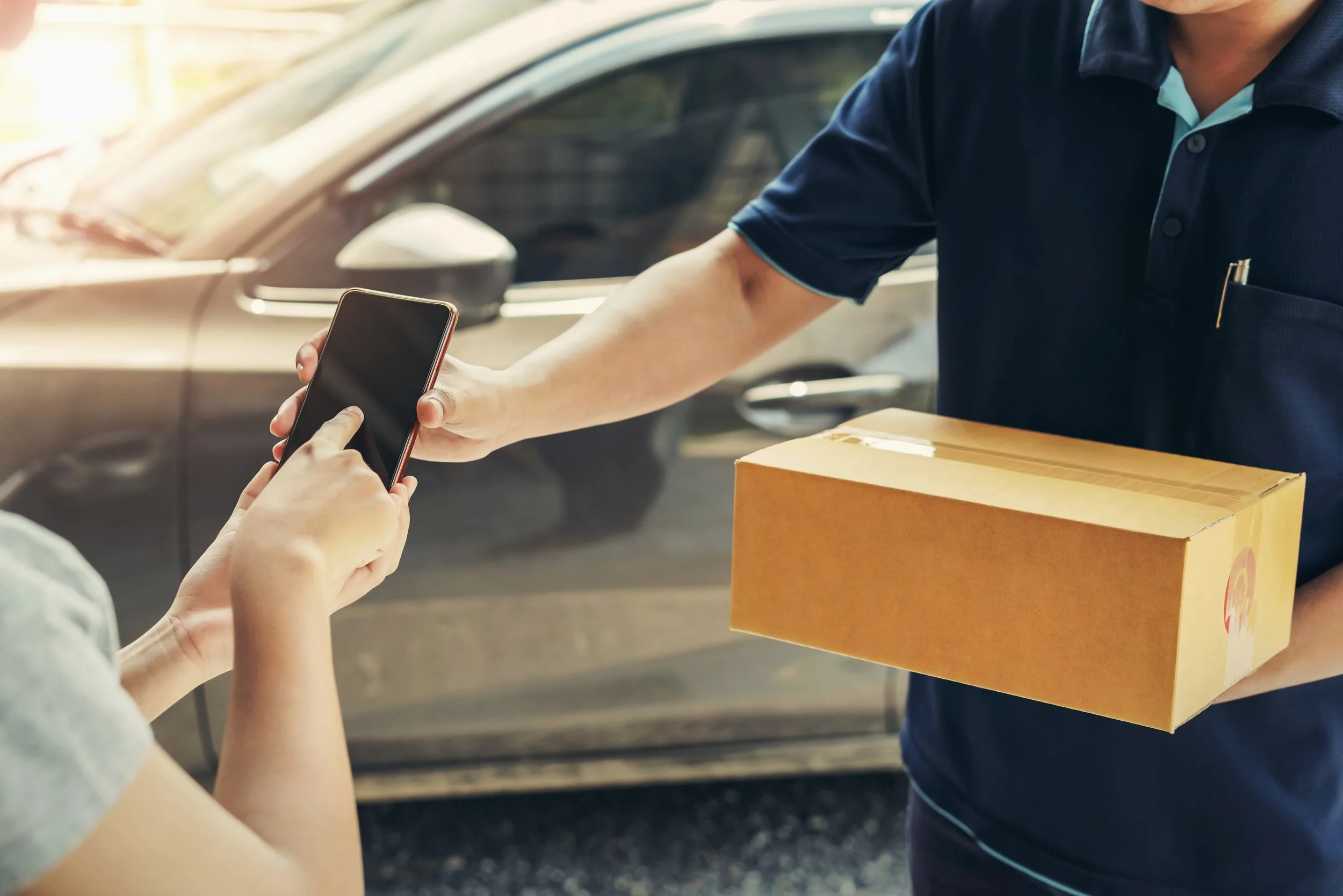 A person is handing a smartphone to another person who is holding a box, with a car in the background.