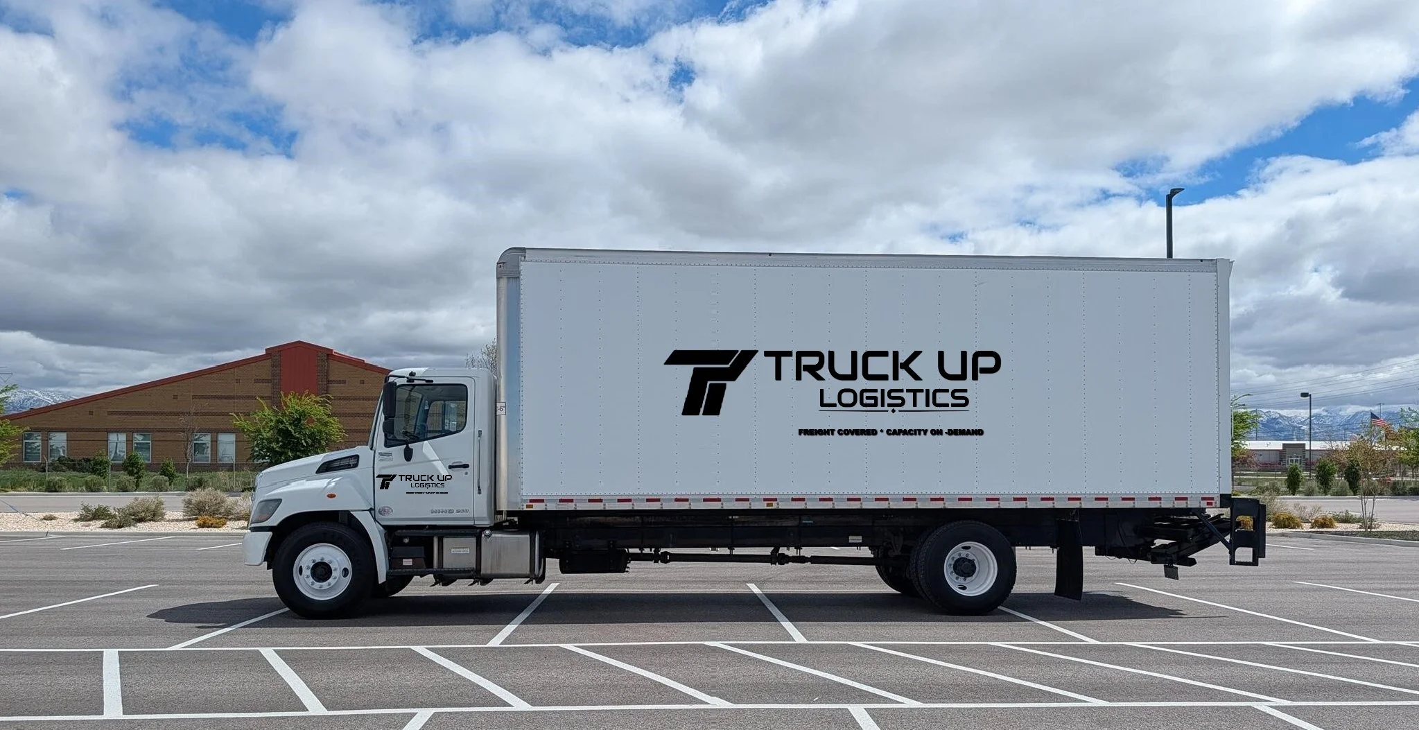 White box truck with a logo of a stylized truck and the words 'Truck Up Logistics' on the side, parked on a lot with other trucks under a clear blue sky.