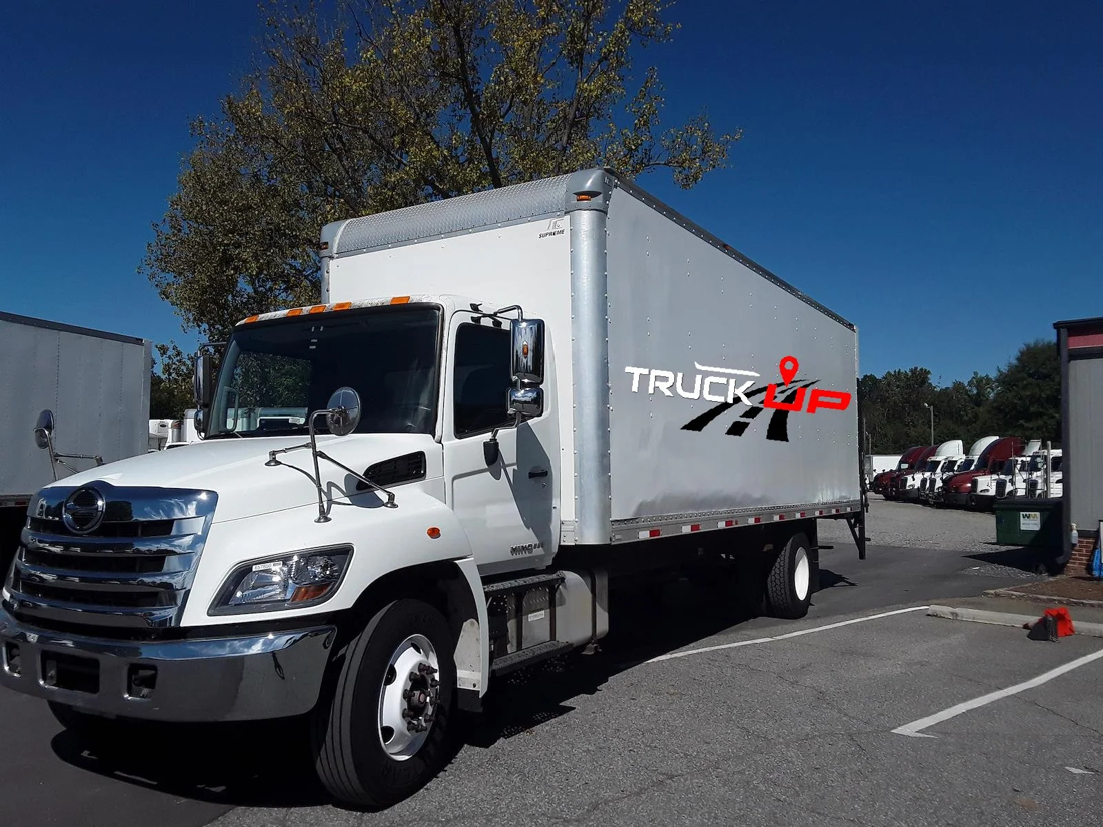 White box truck with a logo reading 'TRUCK UP' parked in a lot, with other trucks visible in the background under a clear blue sky.