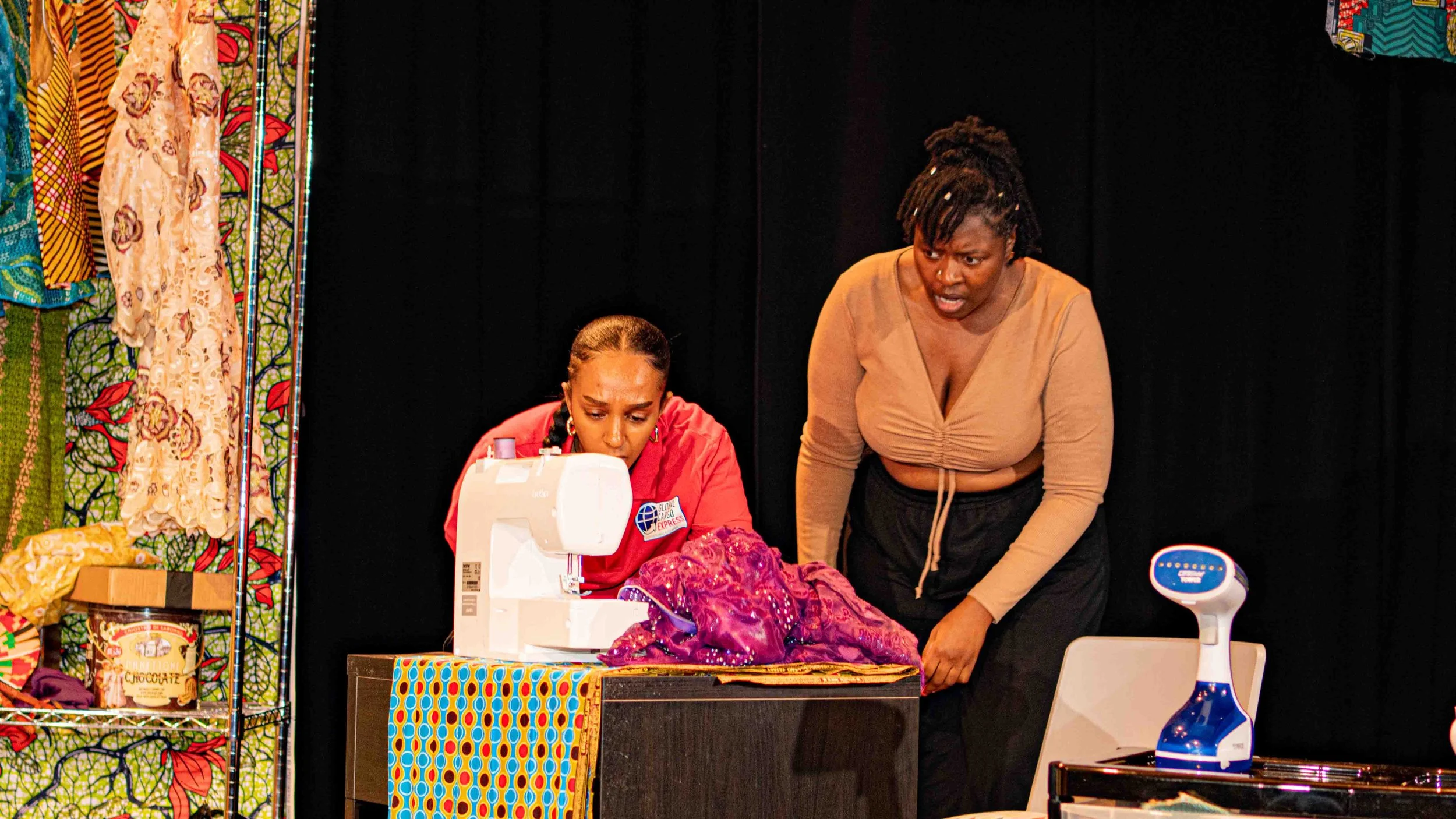 Two women are engaged in a sewing activity on a stage in a theatrical performance with colourful fabric and clothing display in the background. One woman in red is focused on the sewing machine, while the other woman leans over to observe or assist. 
