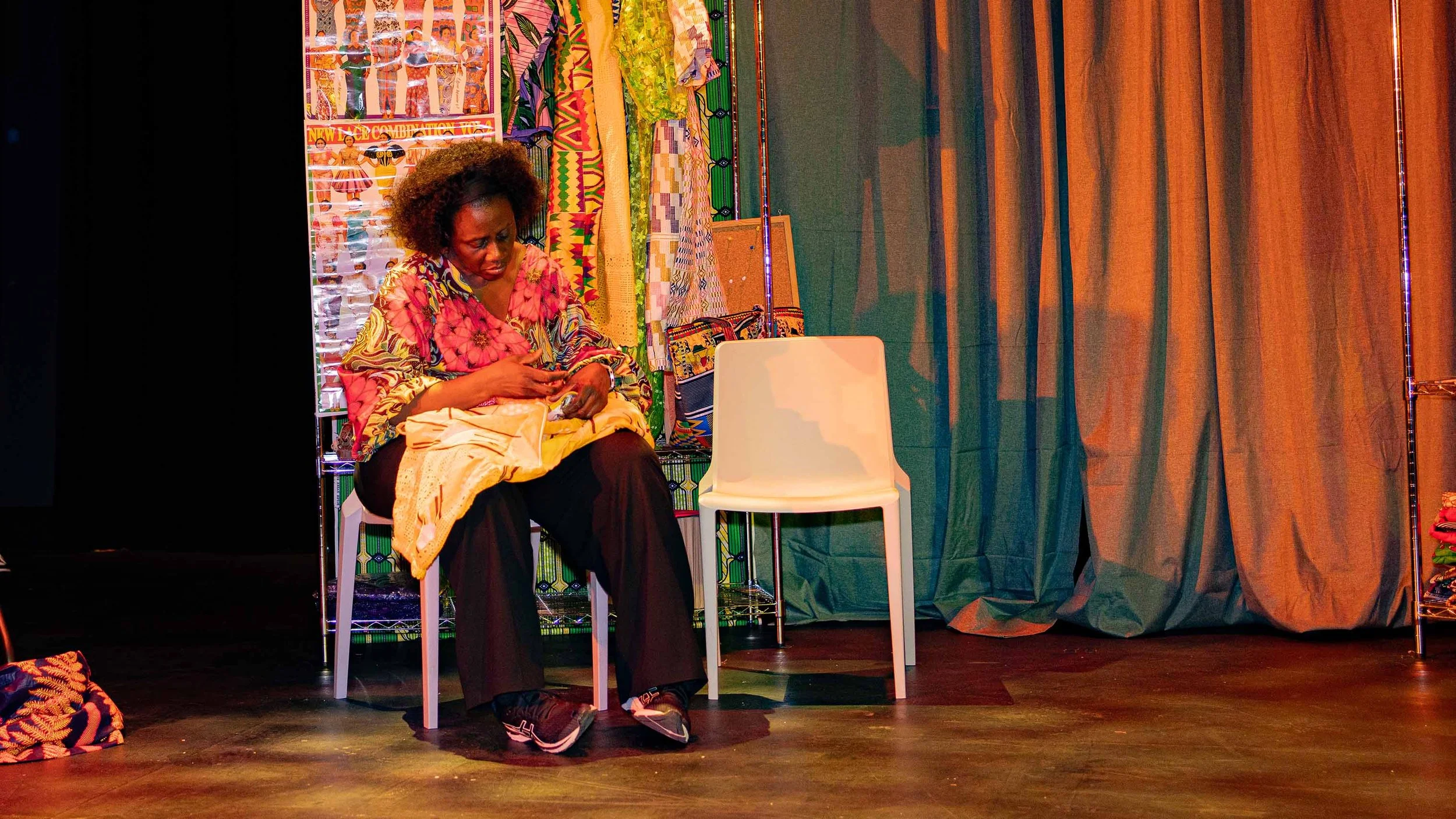 A woman on stage in a theatrical scene, with curly hair sitting on a white chair in front of a colorful fabric display, looking at her phone. There is a small white empty chair next to her and a pile of patterned fabrics on the floor.