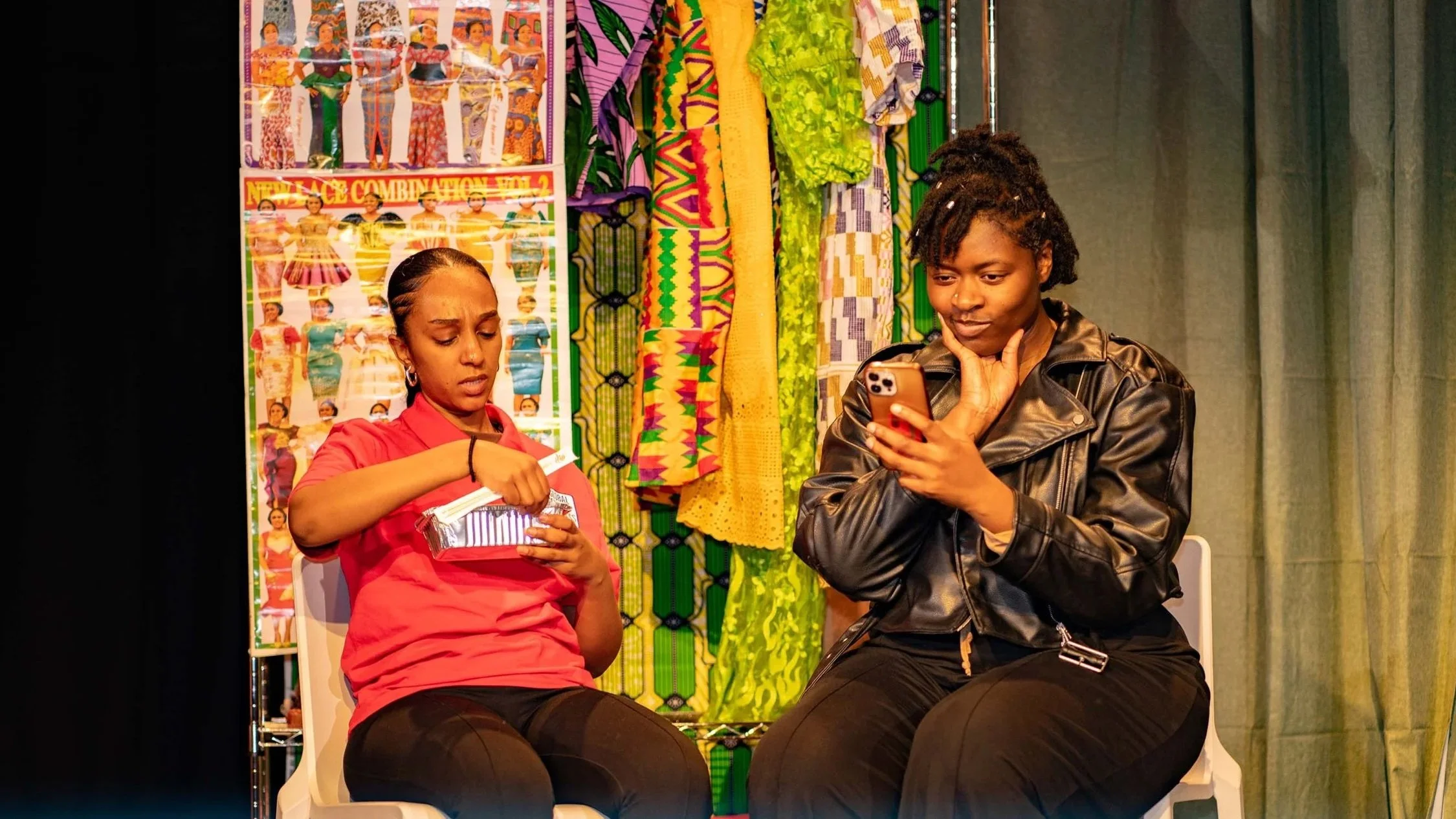 Two women sitting on stage in a theatrical scene, one looking at a phone, the other eating a snack, with colourful fabrics and posters of traditional clothing in the background.