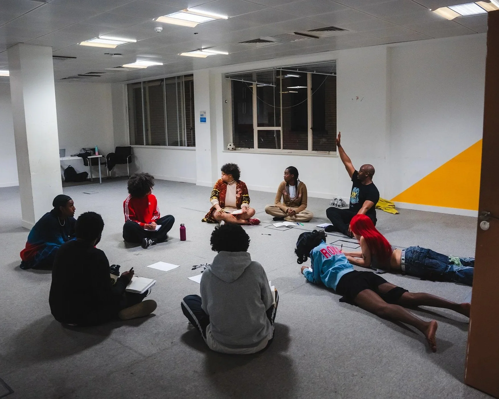 Group of people participating in creative workshop discussion. Some are sitting cross-legged, one person is lying on the floor with red hair, and another person on the floor is raising their hand.