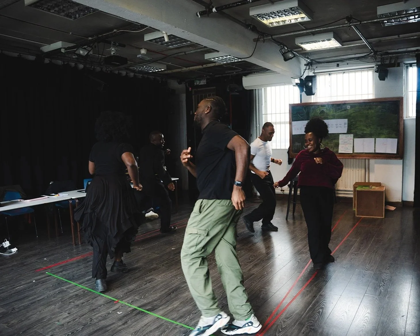 Group of five people dancing and enjoying in a rehearsal studio with black walls, wooden floor, and large windows.
