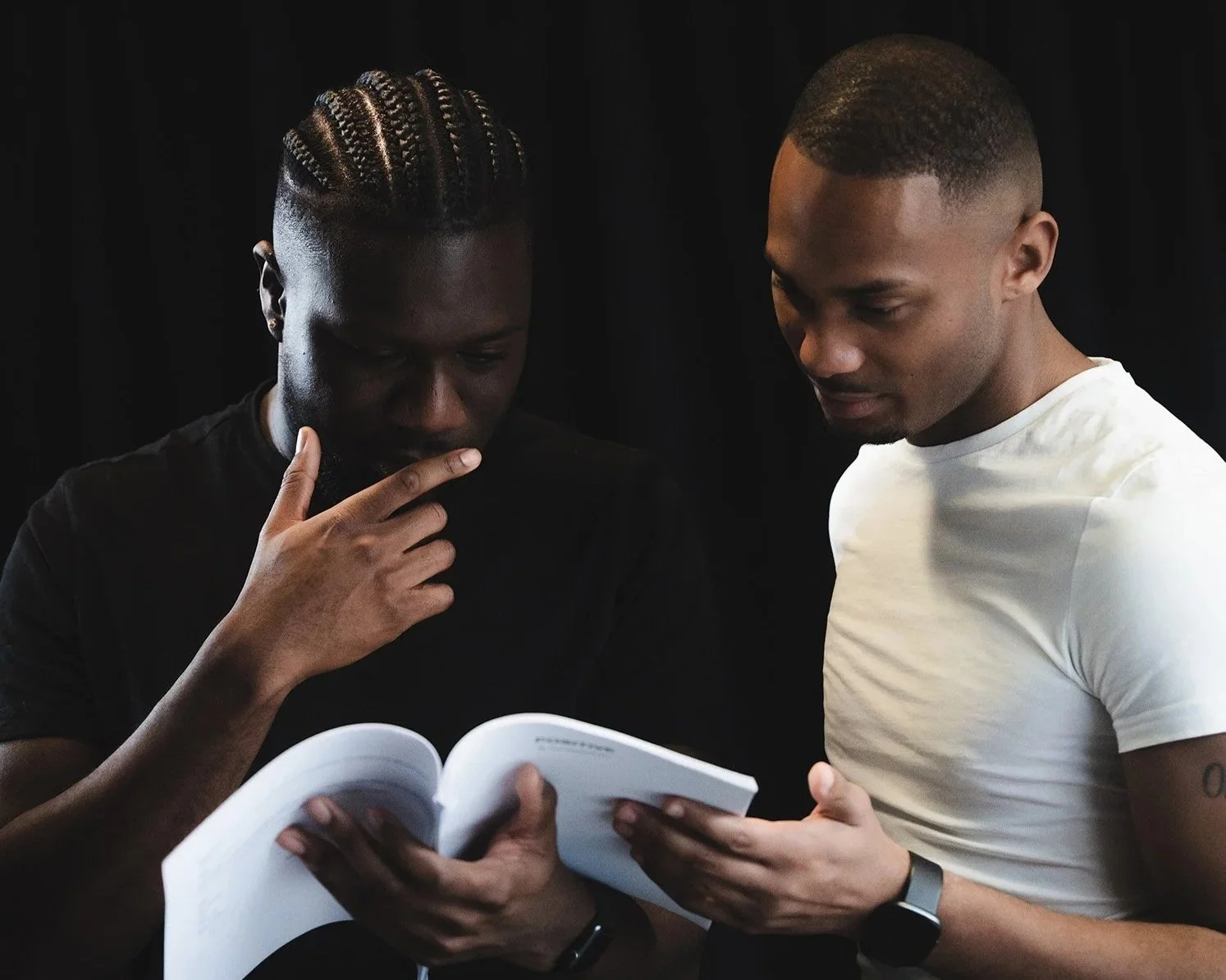 Two men are looking at a script together against a black background. One man has braided hair and is wearing a black shirt, while the other has short hair and is wearing a white shirt and a smartwatch.